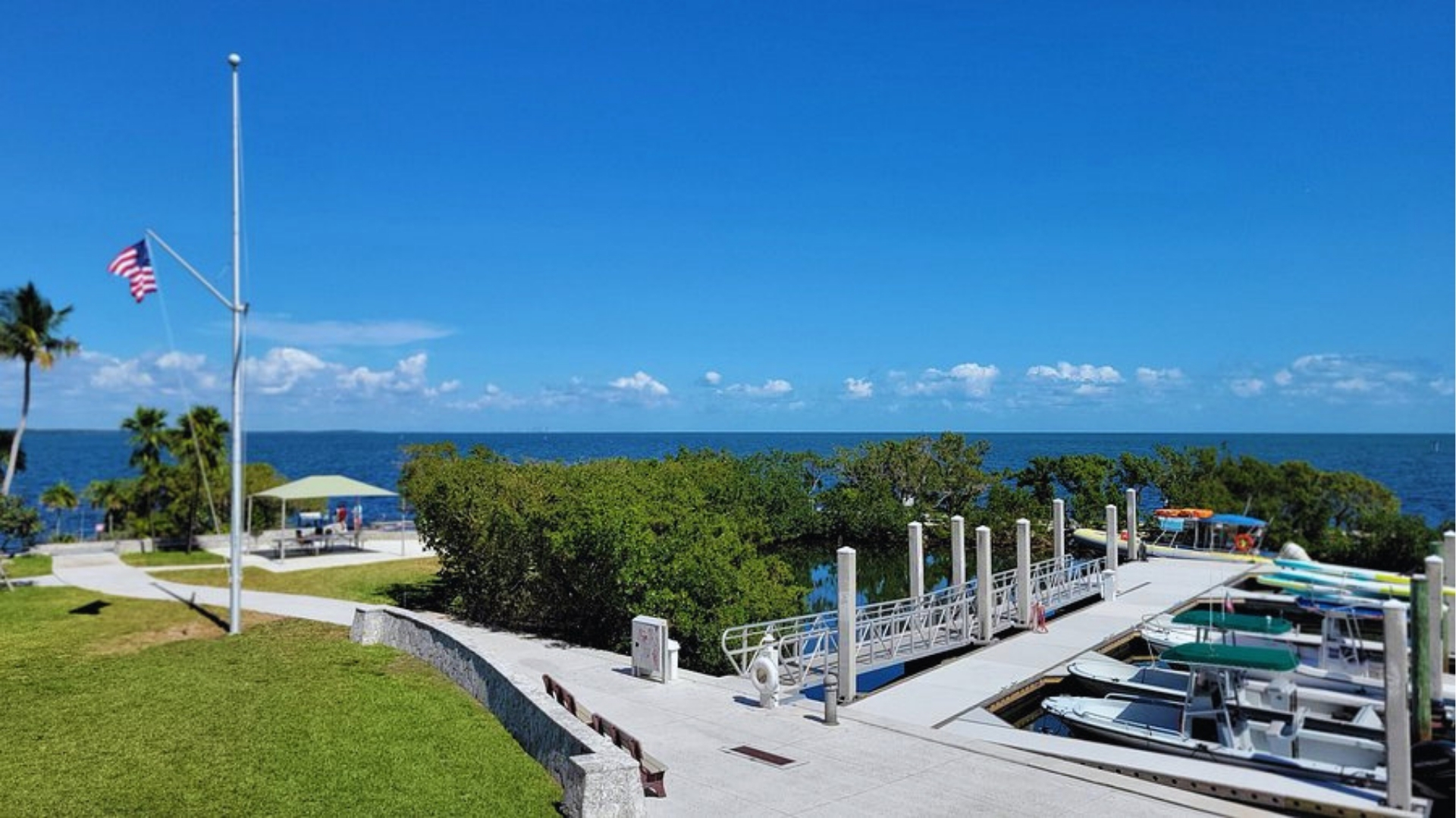 Coastal scene with a dock and boats on the right, an American flag on the left, and a clear blue sky and ocean in the background. Serene and sunny.