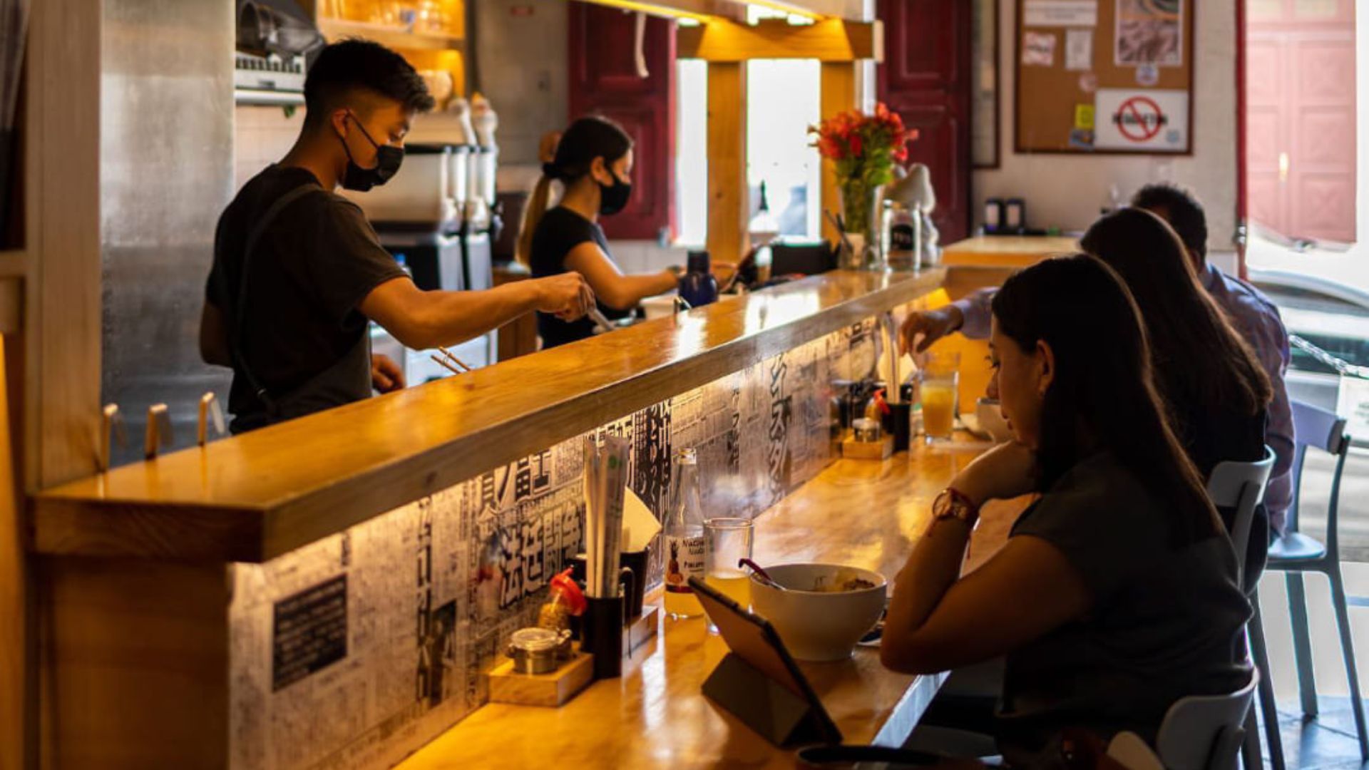 A warm, cozy cafe with a wooden counter where customers sit, enjoying meals. Two masked baristas work behind the counter, creating a lively, inviting atmosphere.