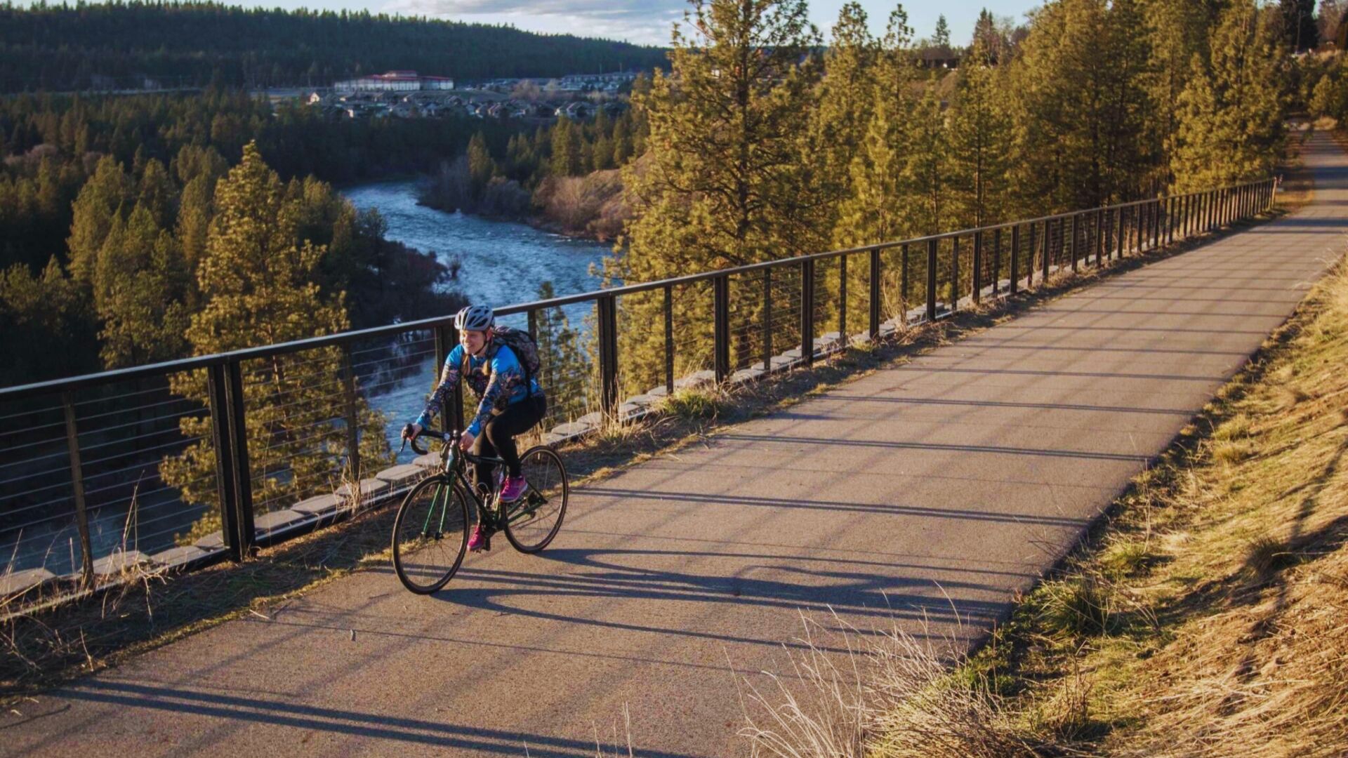A person cycling on a paved path alongside a river, surrounded by greenery and a clear blue sky.