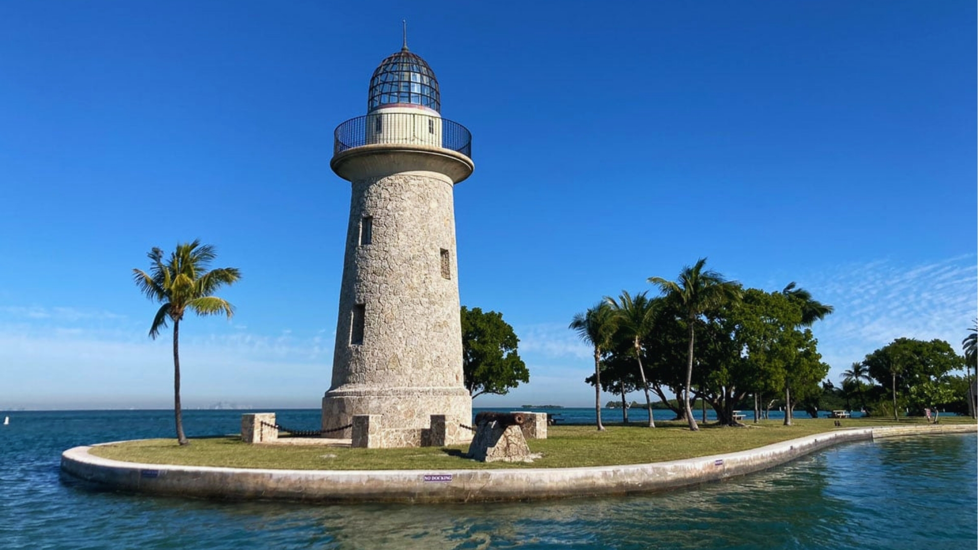 A stone lighthouse with a metal dome stands on a small islet surrounded by turquoise water. Palm trees and a clear blue sky create a serene scene.