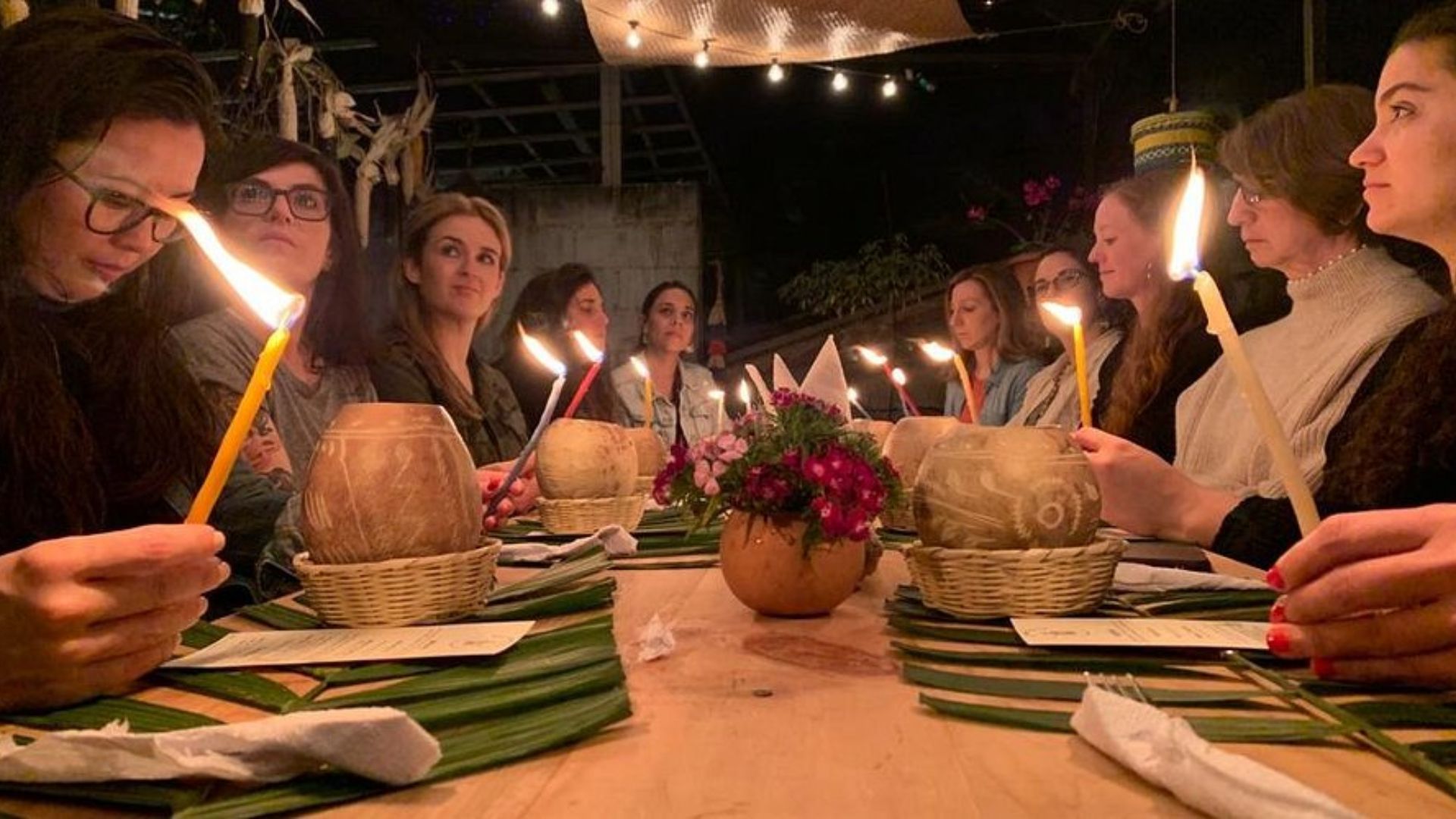 A group of women sits around a wooden table lit by candles in a cozy, dimly lit setting. They hold candles, creating a warm and serene atmosphere.