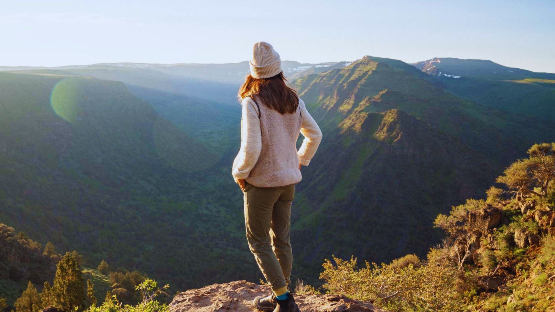 A woman stands on a mountain peak, gazing out over a vast, scenic valley below her.