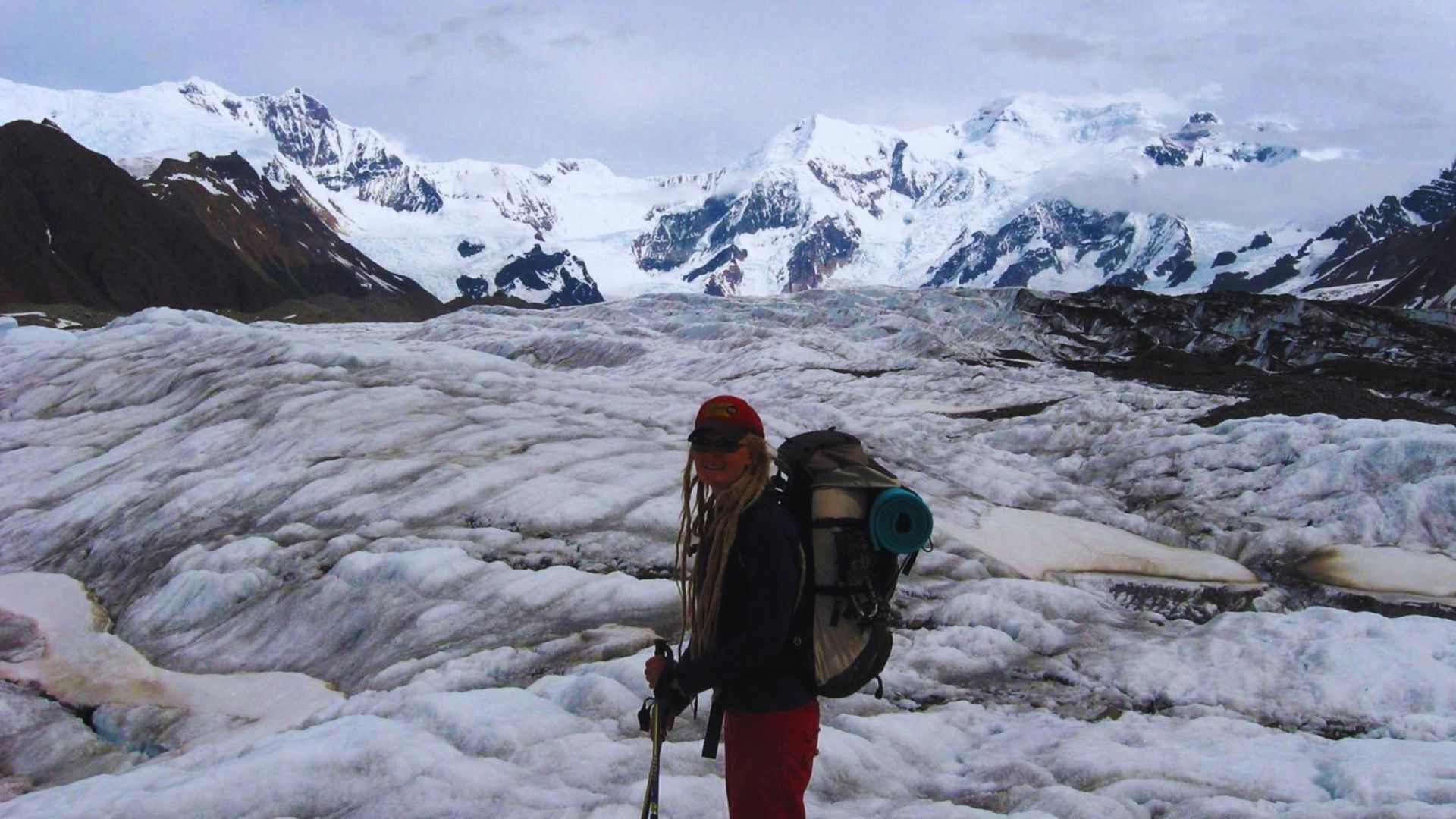 A hiker with a backpack and poles stands on a snow-covered glacier, surrounded by rugged, snowy mountains under an overcast sky, conveying adventure and solitude.