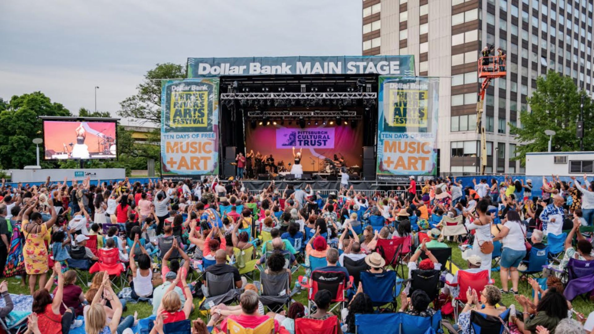 Outdoor music festival with a large crowd seated on the grass, cheering. A band performs on the "Dollar Bank Main Stage." The atmosphere is lively and festive.