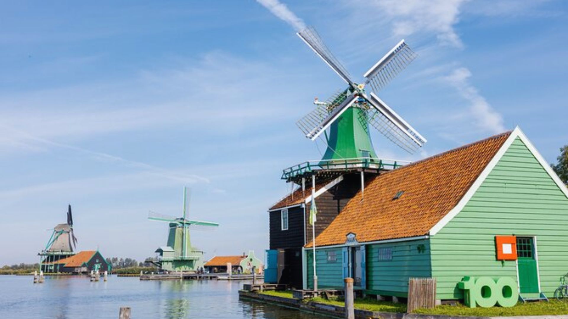 A serene landscape of a Dutch village with traditional green wooden windmills and houses beside a calm waterway under a clear blue sky.