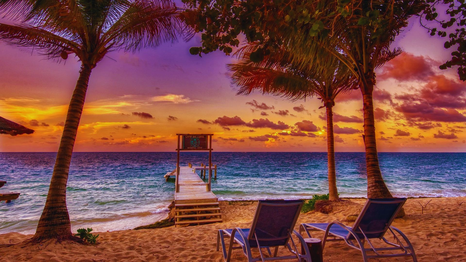 Sunset beach scene with a vibrant sky of orange and purple hues. Palm trees frame a wooden pier leading to the ocean, with two empty lounge chairs on the sandy shore, creating a peaceful, tropical ambiance.