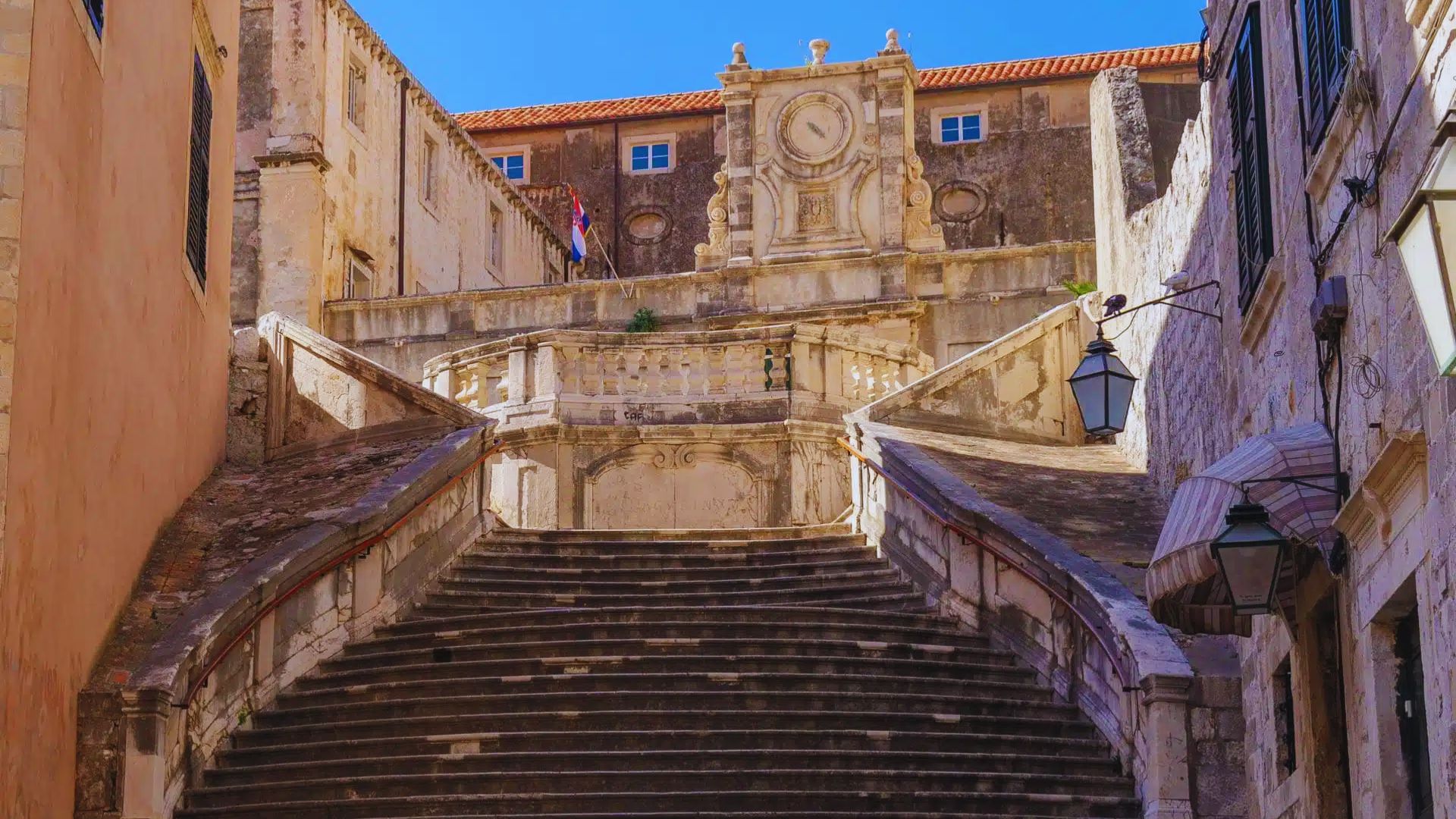 Stone staircase leads up to an ornate clock tower on a sunny day. Rustic buildings flank the stairs, creating a historic and grand atmosphere.