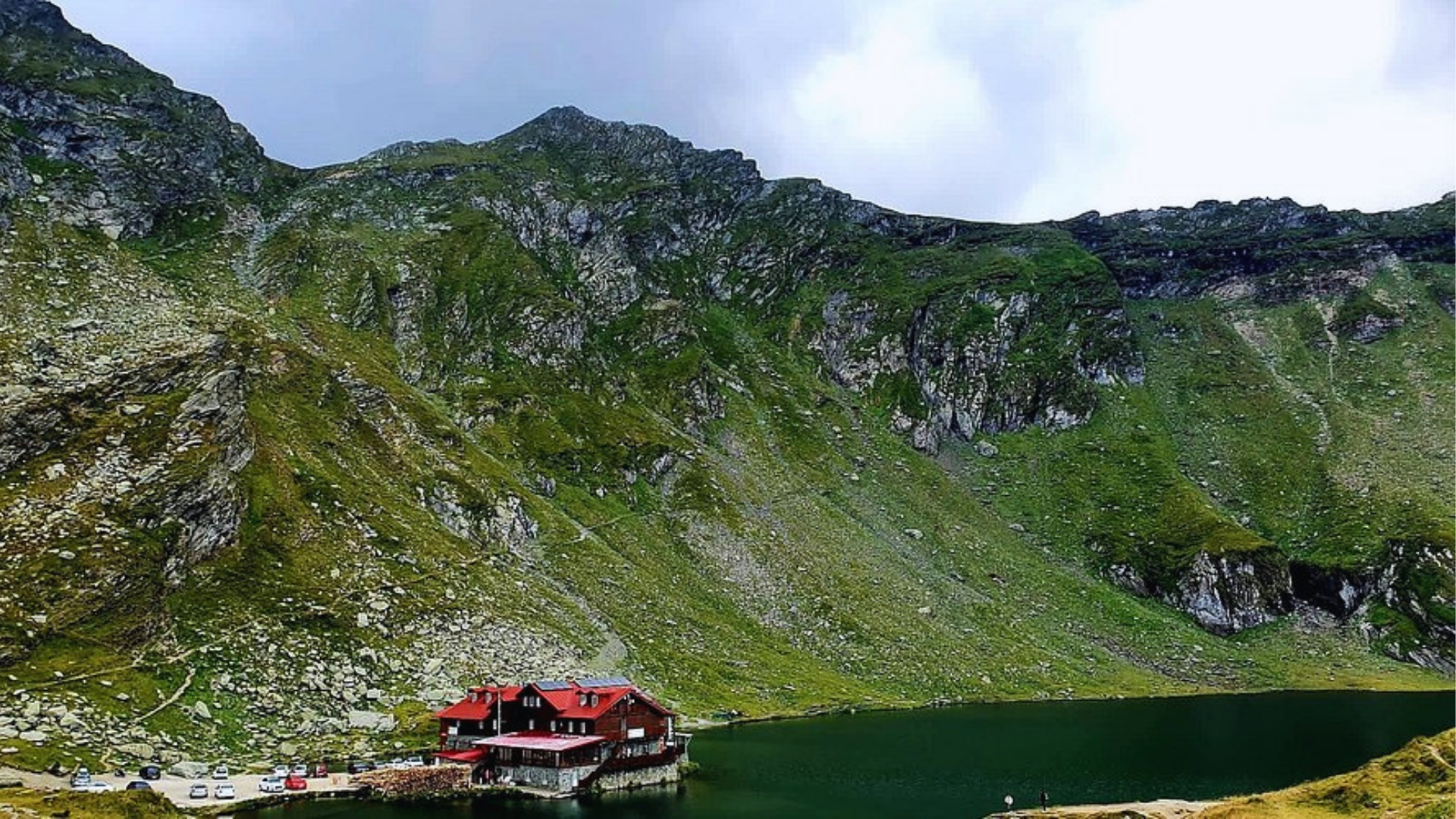 Mountain landscape with a red-roofed cabin by a tranquil lake, surrounded by steep, rocky green hills under a cloudy sky. Peaceful and remote atmosphere.