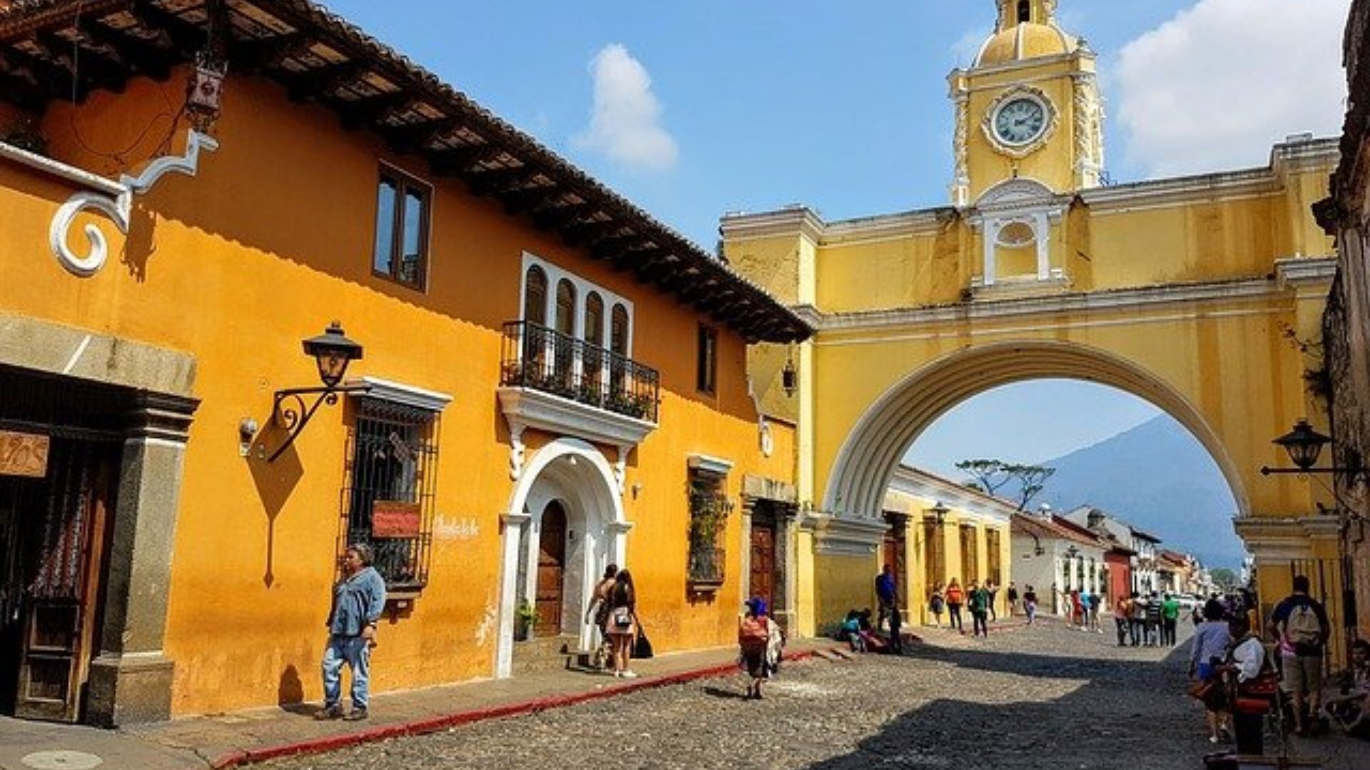 A vibrant street scene in Antigua, Guatemala, featuring a yellow clock tower arch, colorful colonial buildings, cobblestone road, and people strolling.