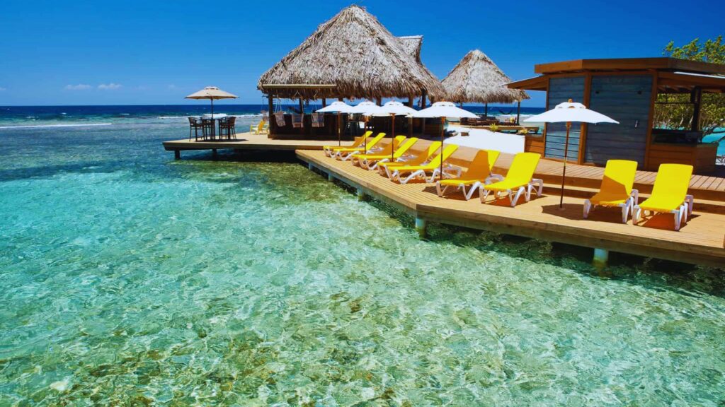 Tropical beach scene with clear turquoise water, yellow lounge chairs, and white umbrellas on a wooden deck. Thatched-roof huts convey a sunny, relaxing vibe.