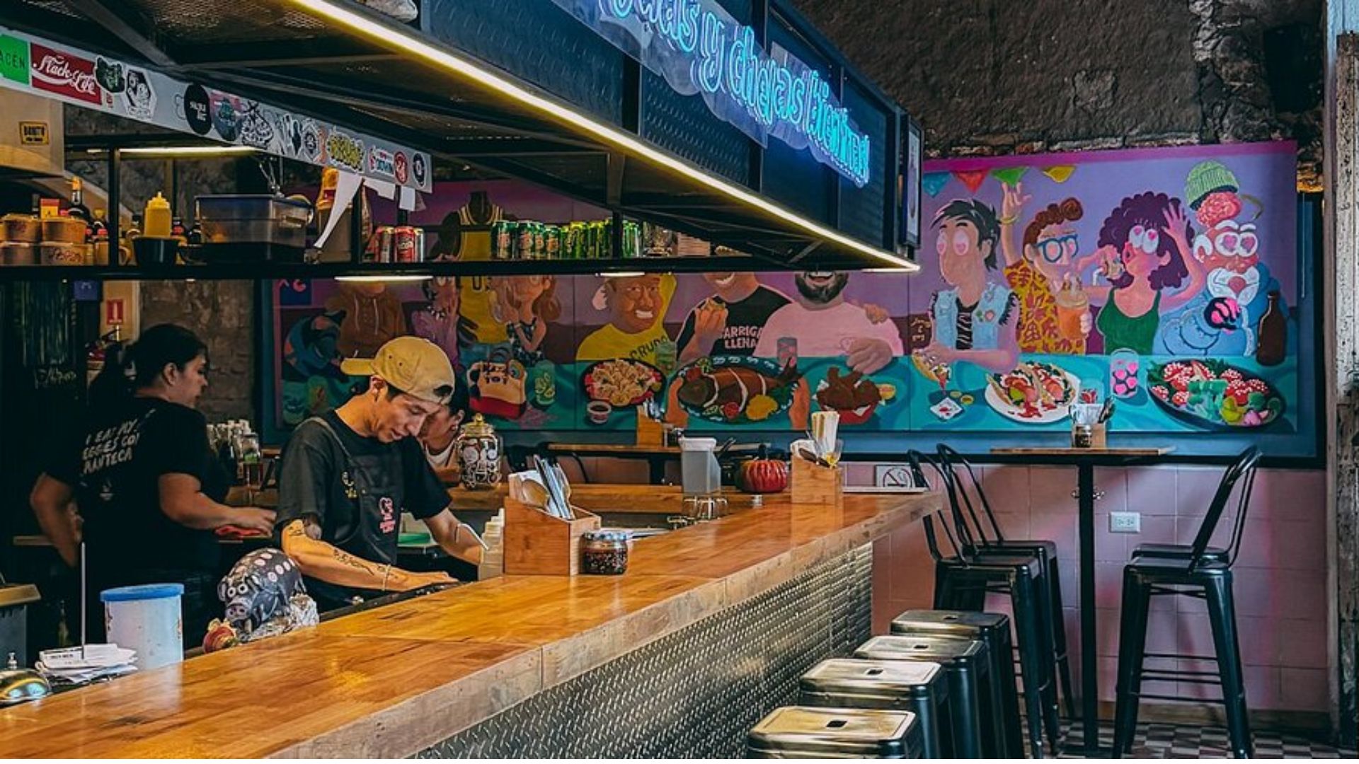 A lively taco bar with a vibrant mural depicting joyful diners enjoying tacos. The bartender is working behind a wooden counter with stools. Warm, inviting atmosphere.