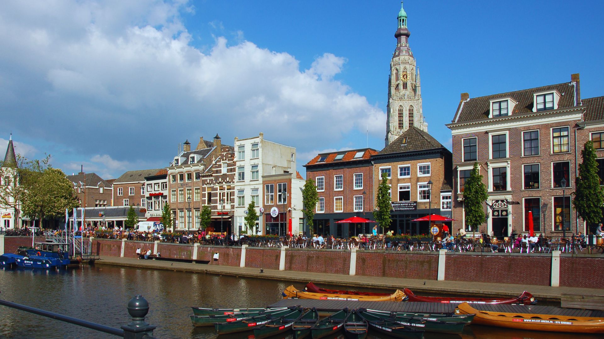 Canal-side scene with colorful historic buildings in Breda, Netherlands. A church tower rises in the background; small boats are docked in the water.