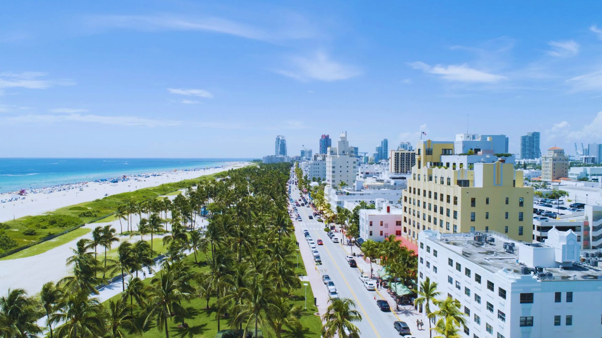 Aerial view of Miami Beach with palm-lined roads, white sandy beach, blue ocean, and art deco buildings. Clear sky adds to the vibrant, sunny atmosphere.