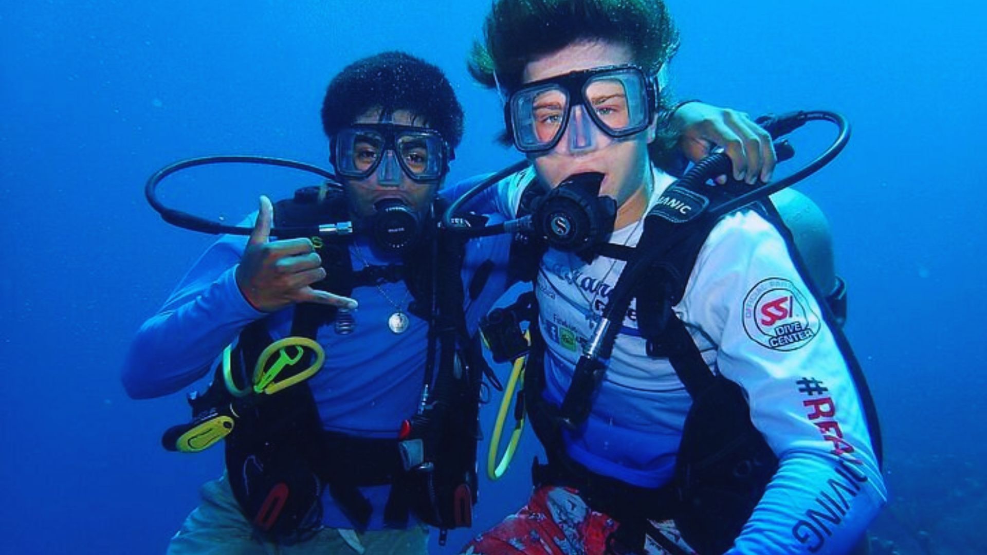 Two scuba divers underwater, wearing masks and gear, looking at the camera. One diver gestures with a hand sign. The scene conveys adventure and excitement.
