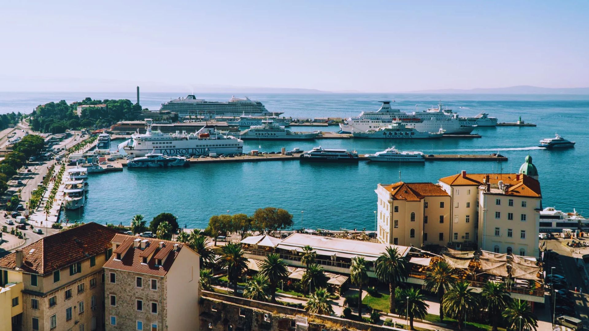 Aerial view of a bustling harbor in Split, Croatia, with several large cruise ships docked. The foreground features historic buildings and lush palm trees.