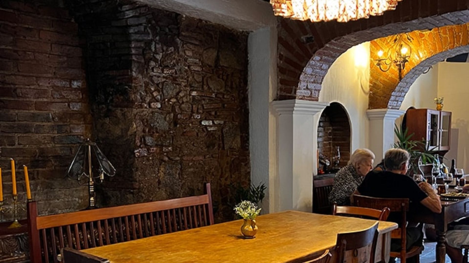 A cozy restaurant interior with rustic brick walls and archways. A wooden table is centered, set with candles, while two people dine, creating a warm, intimate atmosphere.