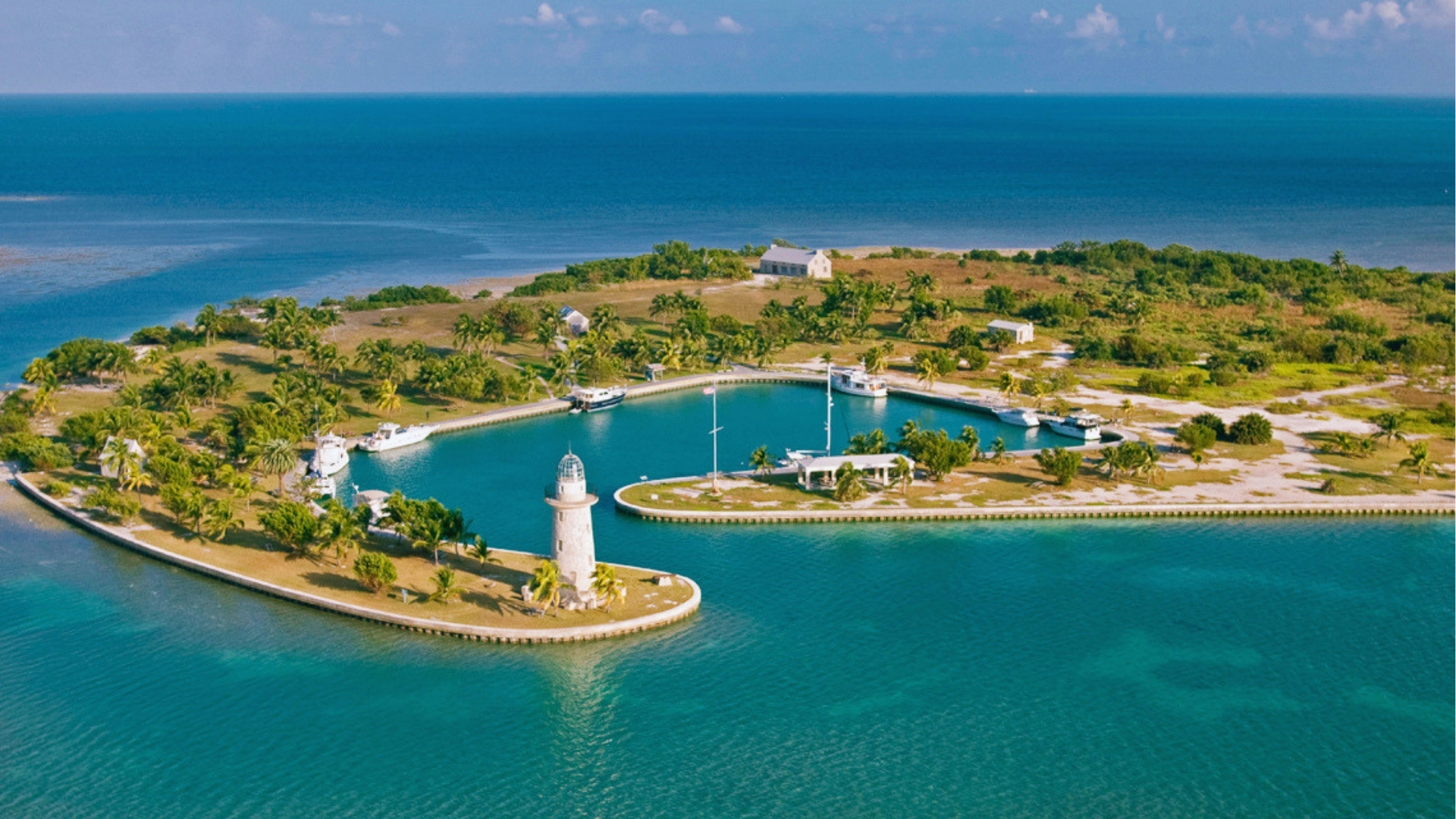Aerial view of a picturesque island with lush greenery and palm trees. A lighthouse and marina are surrounded by calm, turquoise waters under a clear sky.