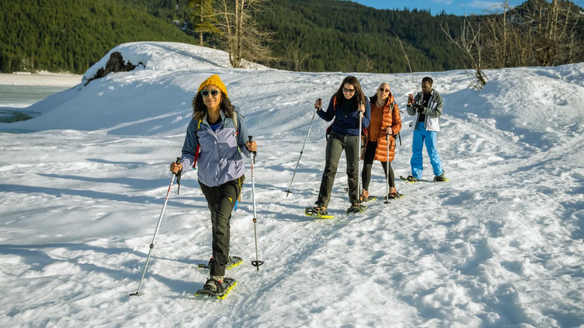 Four people snowshoeing on a sunny day, surrounded by snowy hills and forest. They appear cheerful, enjoying an outdoor winter adventure.