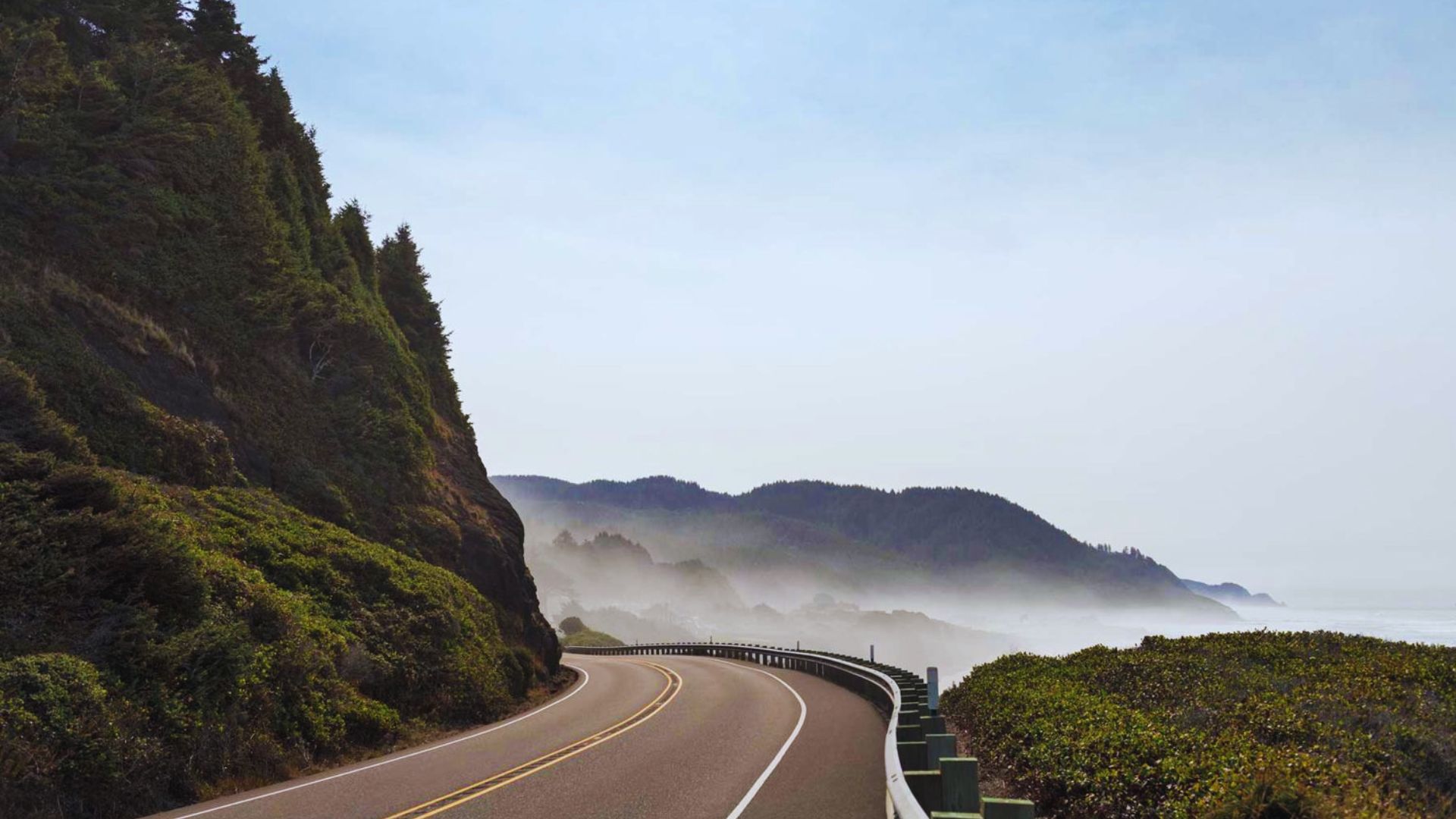 Coastal road curves beside lush cliffs, misty hills, and ocean, under a clear sky, creating a serene, scenic drive.