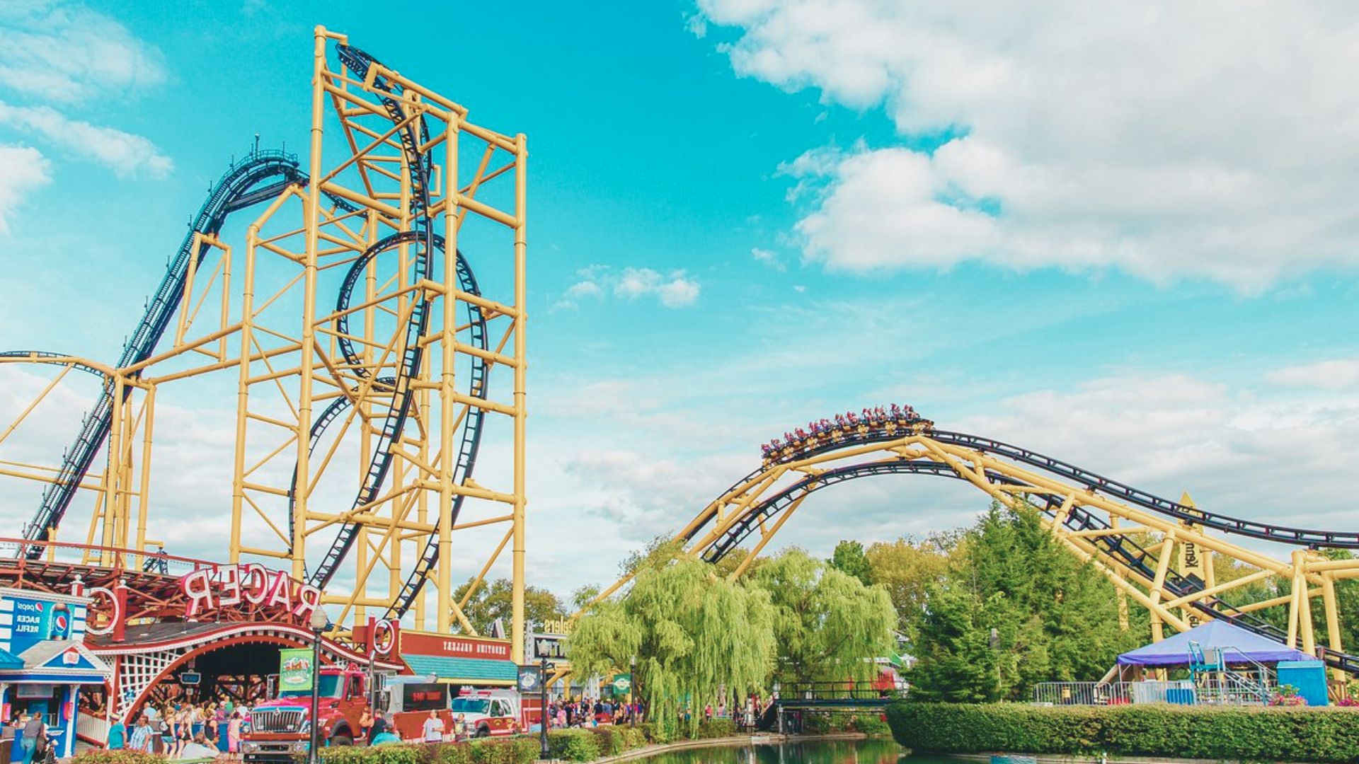 A vibrant amusement park scene with a towering roller coaster under a bright blue sky. Lush greenery surrounds the area, creating a fun and lively atmosphere.