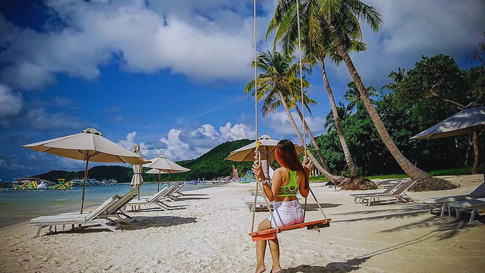 A woman swings on a beach surrounded by palm trees, enjoying a sunny day by the ocean.