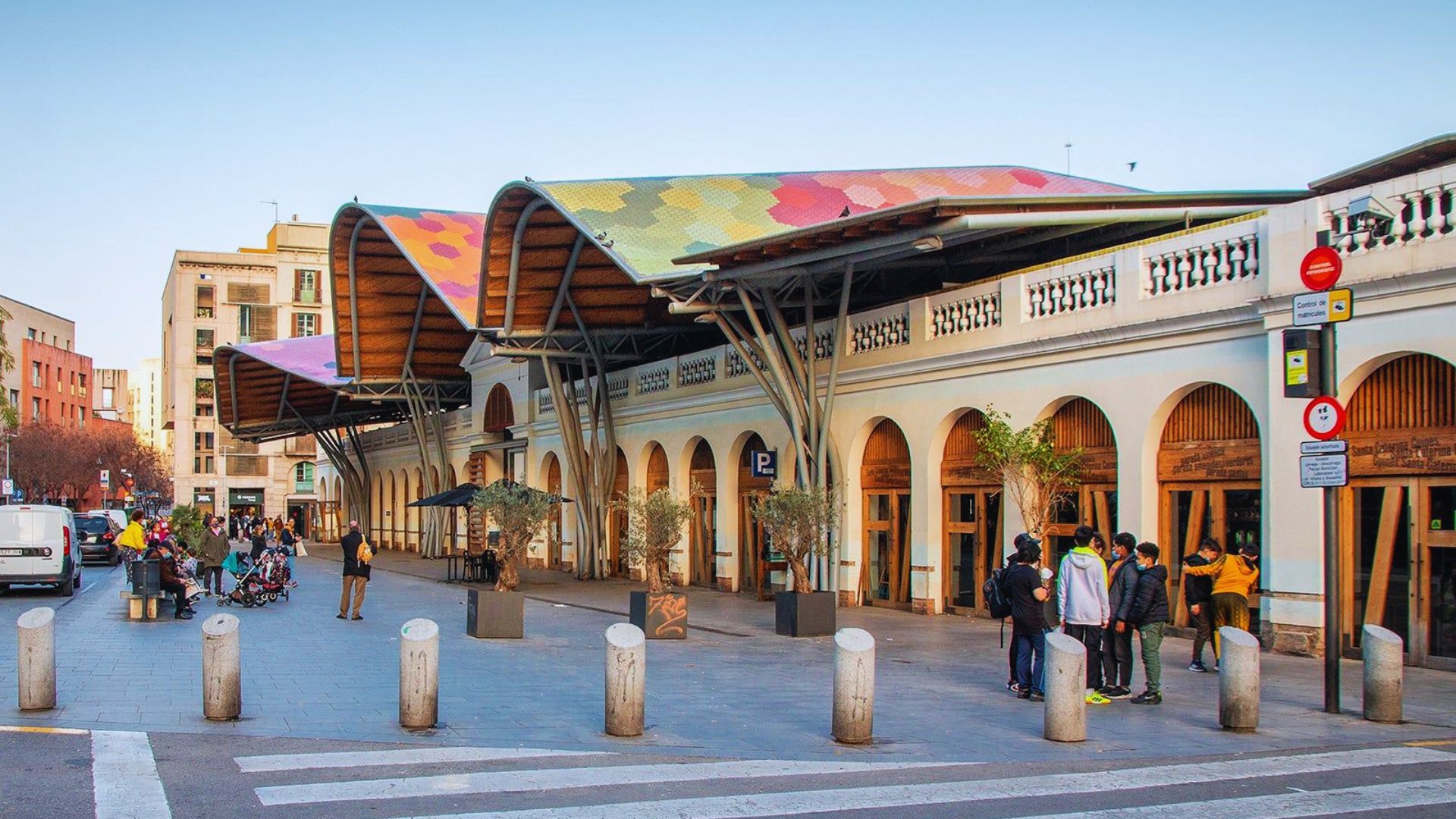 A vibrant market with an undulating, colorful roof resembling mosaic tiles. People gather outside, creating a lively and welcoming atmosphere.