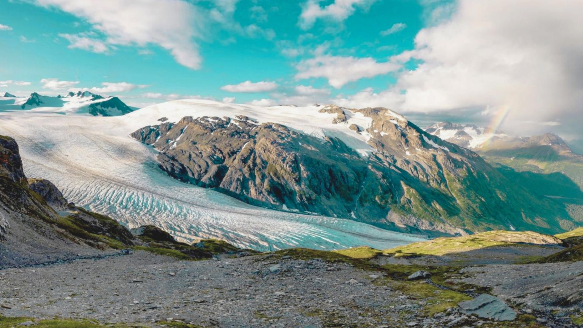 Snow-capped mountains and a glacier under a bright blue sky with scattered clouds. A faint rainbow arcs over lush green valleys, creating a serene landscape.