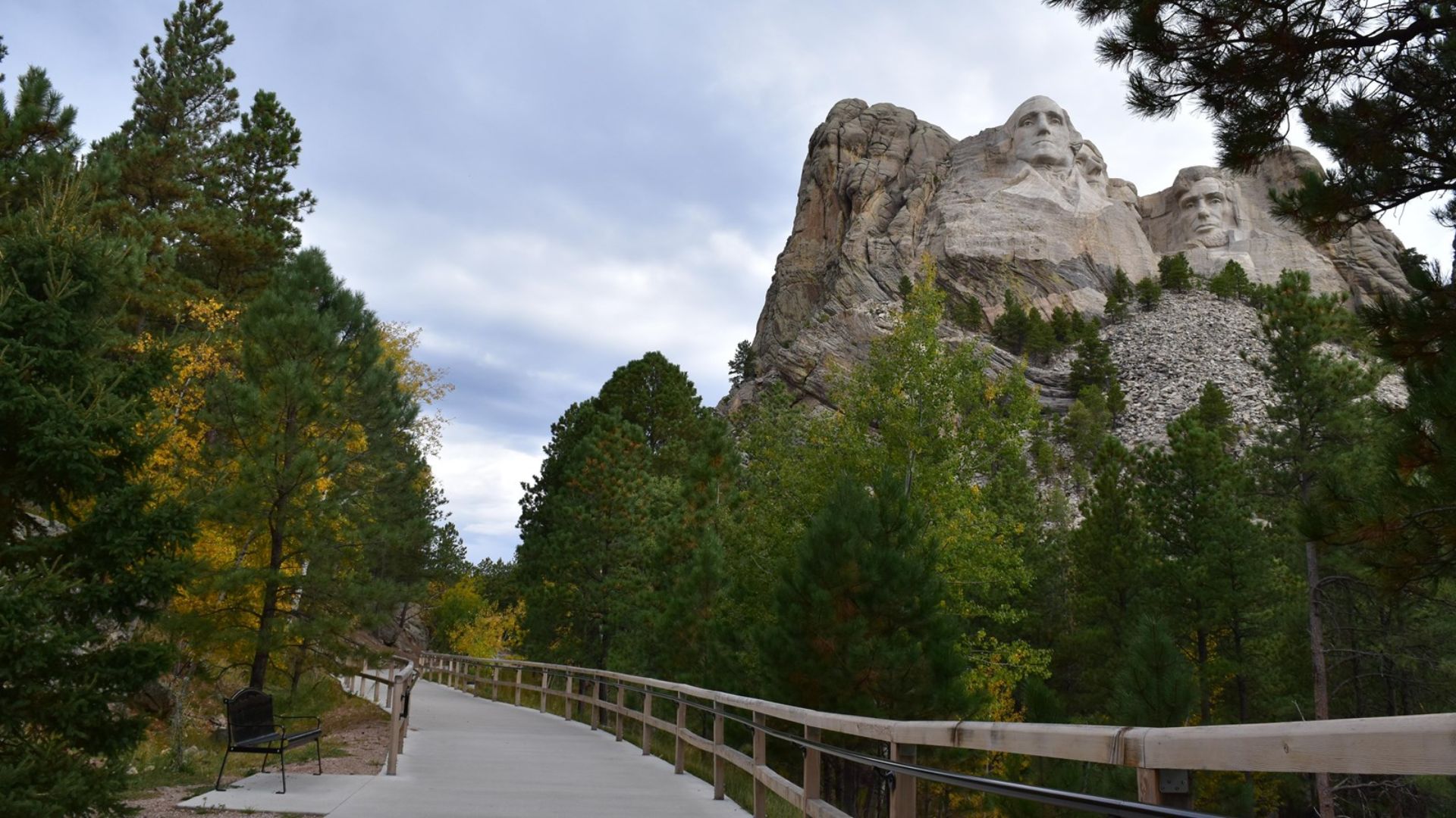 A paved walkway leads through lush green trees to Mount Rushmore, where two presidential faces are carved into the rocky mountainside beneath a cloudy sky.