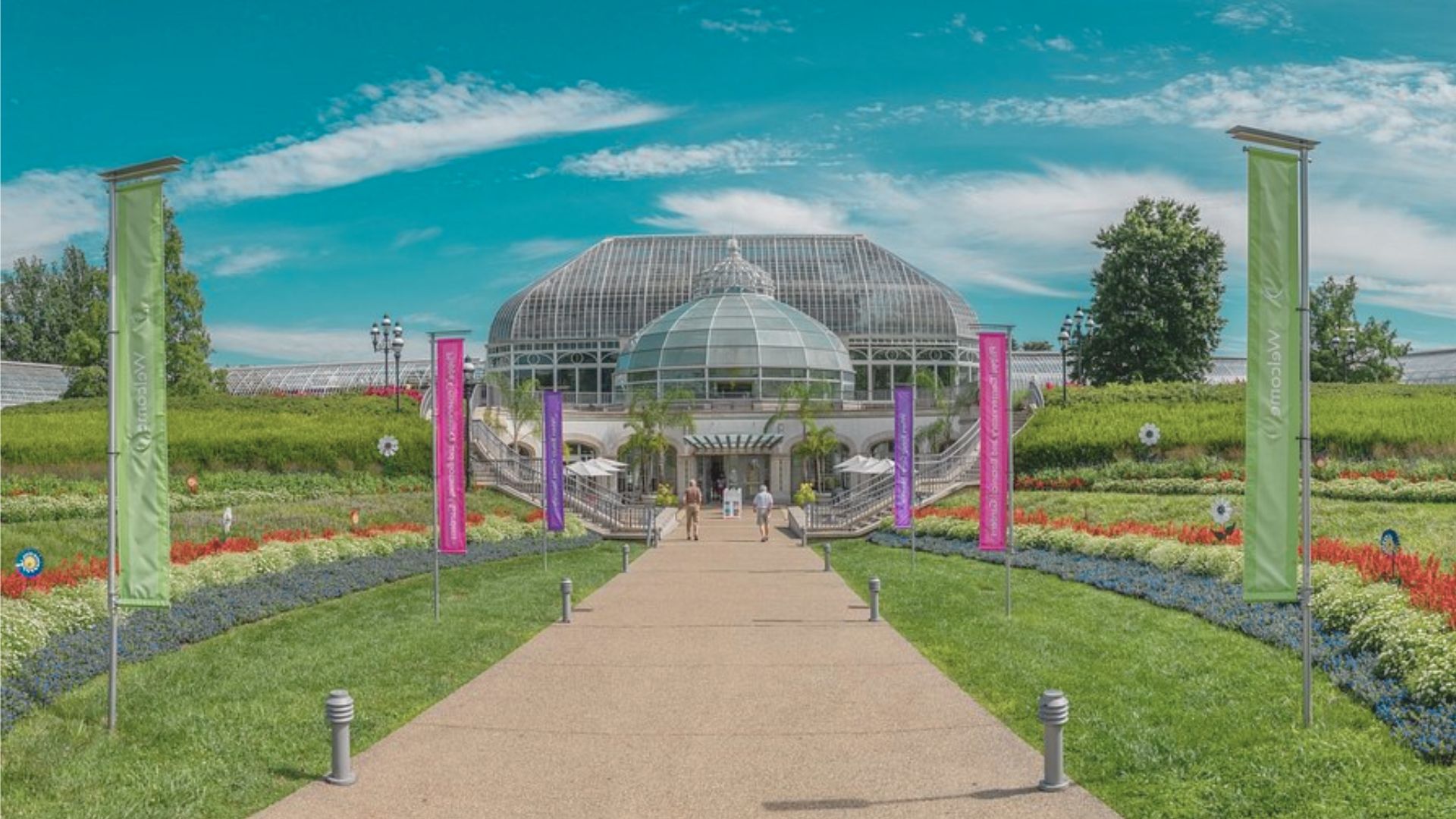 Pathway leading to a glass-domed conservatory surrounded by colorful flowerbeds and tall flags under a bright blue sky with scattered clouds.