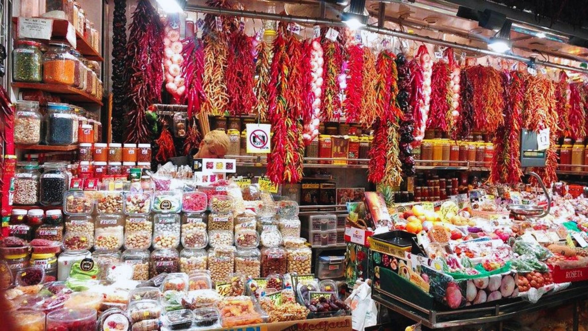 Bustling market stall with vibrant hanging chilies and garlic. Shelves packed with spices, nuts, and jars convey rich scents and a lively atmosphere.