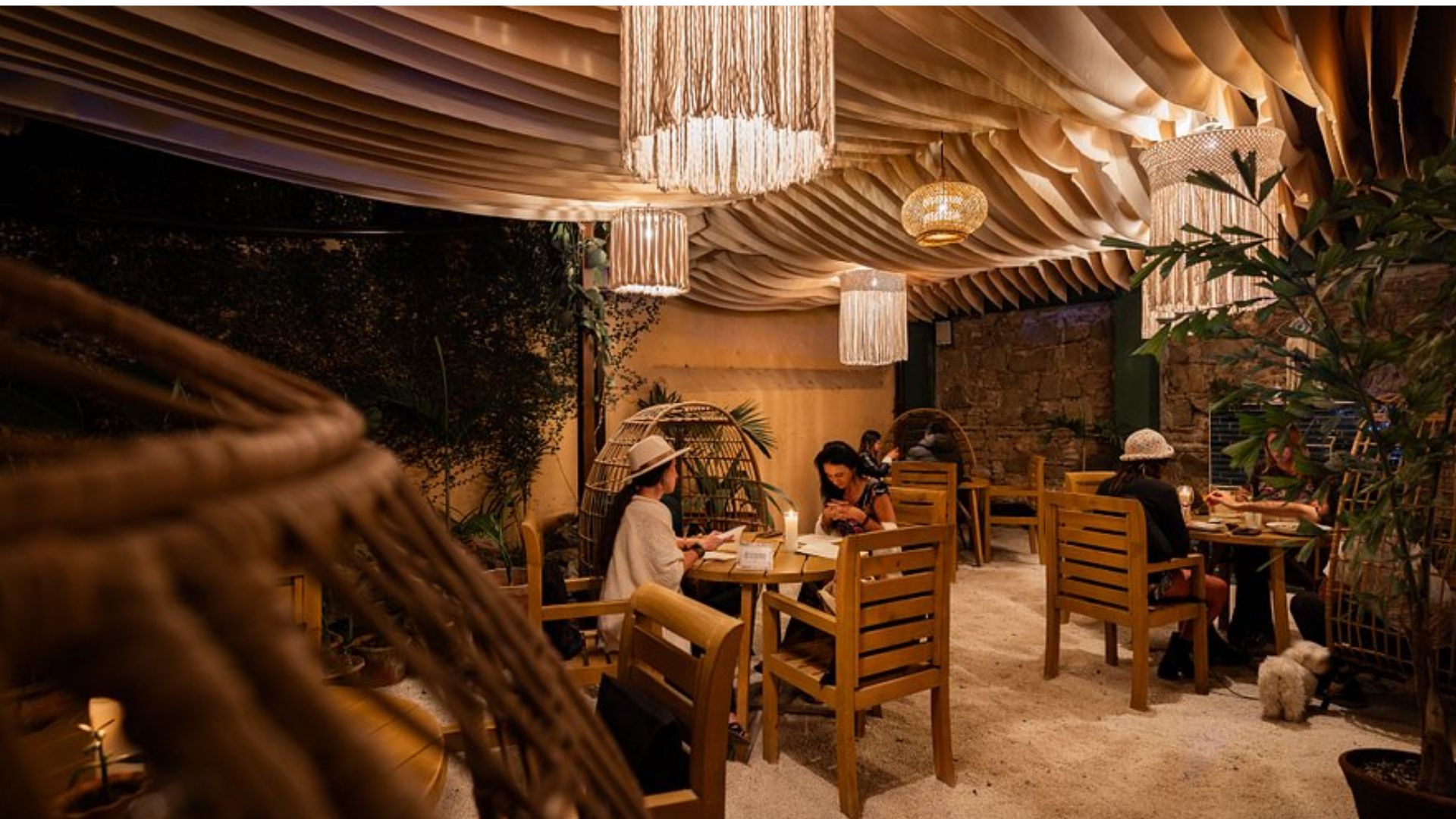 Cozy outdoor café with warm lighting, wooden furniture, and hanging wicker lamps. Guests, wearing hats, chat and enjoy drinks, creating a relaxed atmosphere.