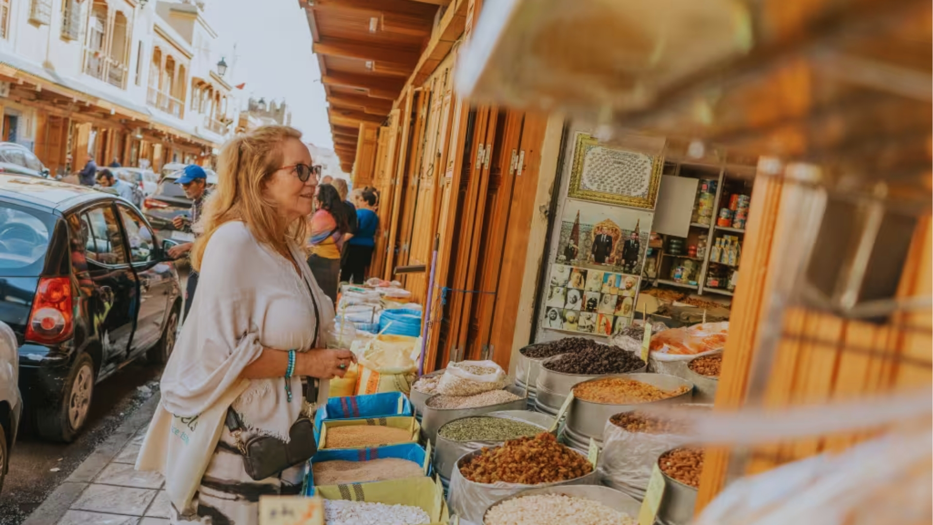 A woman in a light wrap observes assorted spices and grains in open sacks at a vibrant outdoor market. The atmosphere is bustling and colorful.