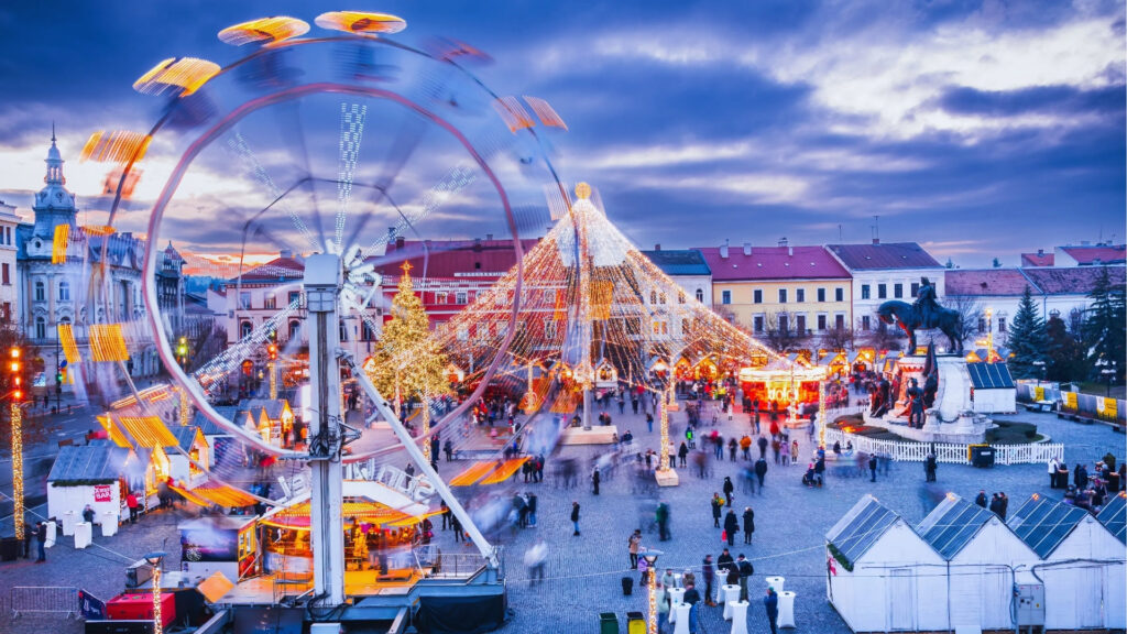 A festive Christmas market scene at dusk with a spinning Ferris wheel, a large Christmas tree adorned with lights, and people walking around, creating a lively atmosphere.