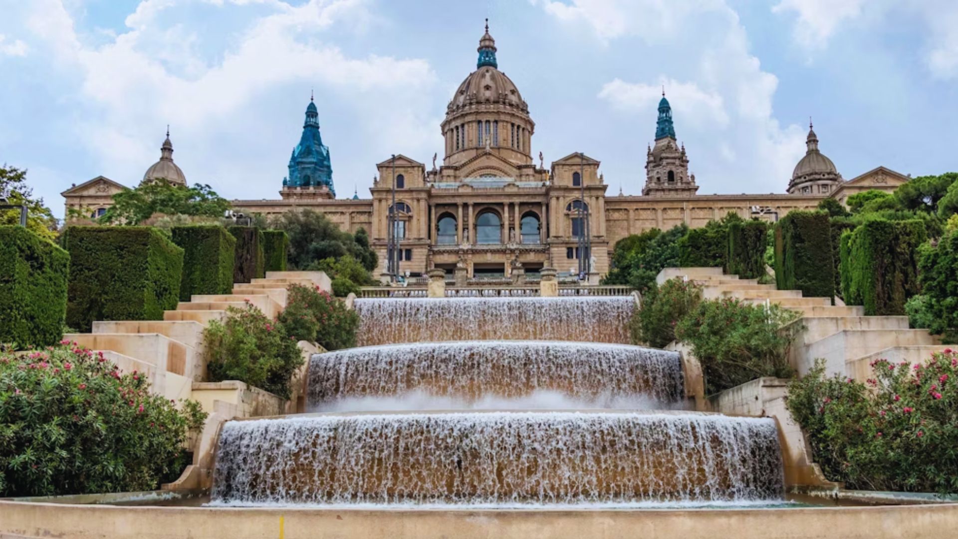 Grand palace with ornate facade and domed towers, surrounded by lush greenery. Cascading water flows in tiered fountains in the foreground.