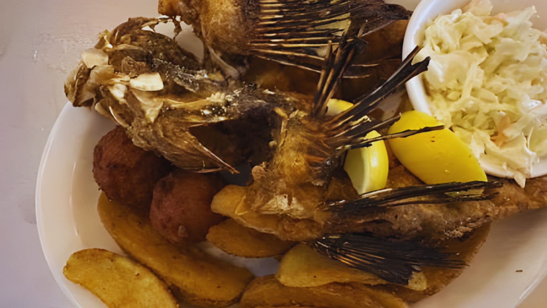 A plate with crispy fried fish, seasoned potato wedges, hush puppies, lemon wedges, and a side of coleslaw in a bowl, creating a rustic, hearty meal.