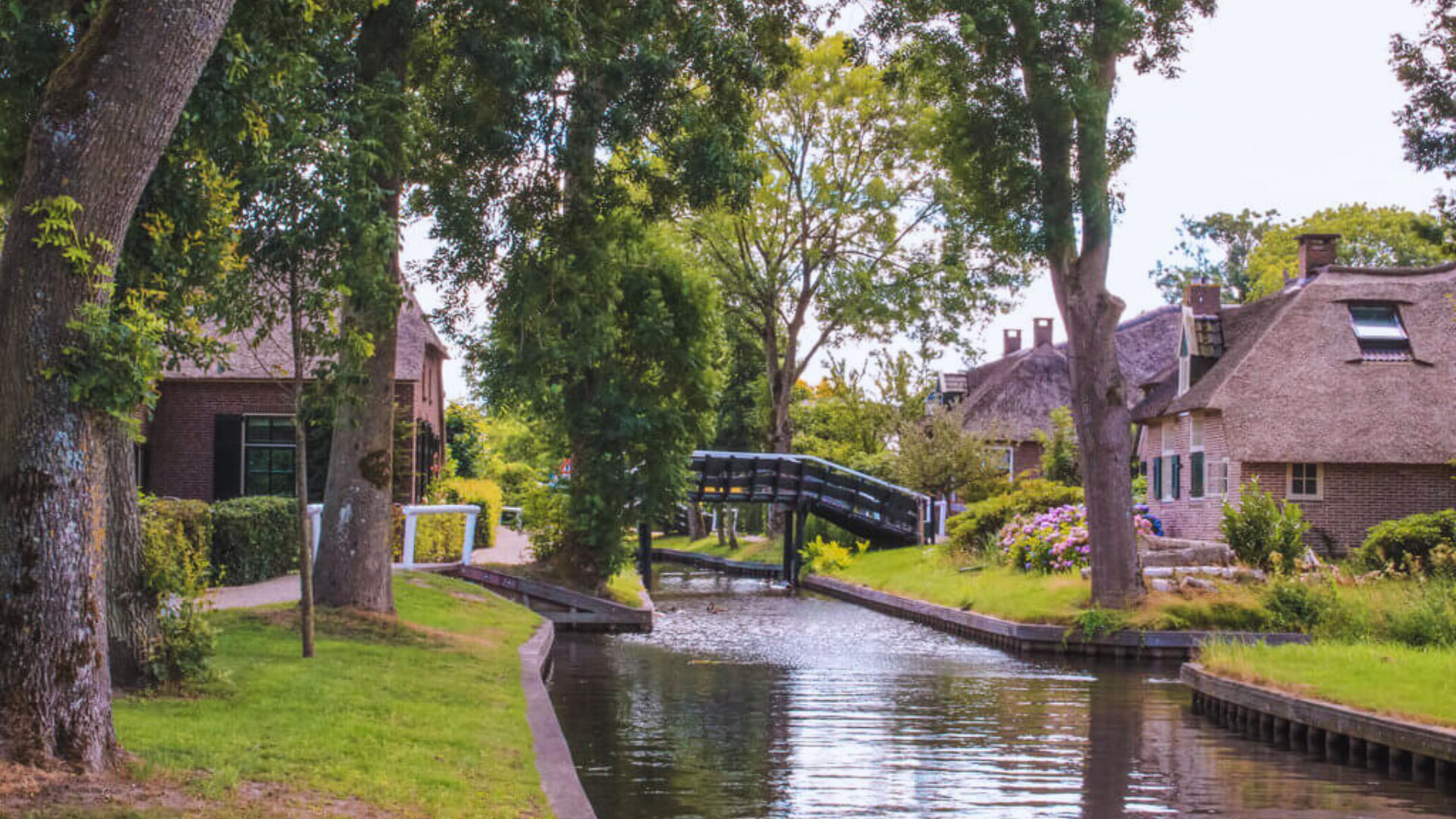 A serene canal scene with lush green trees, charming brick houses with thatched roofs, and a quaint arch bridge, evoking a peaceful, idyllic atmosphere.