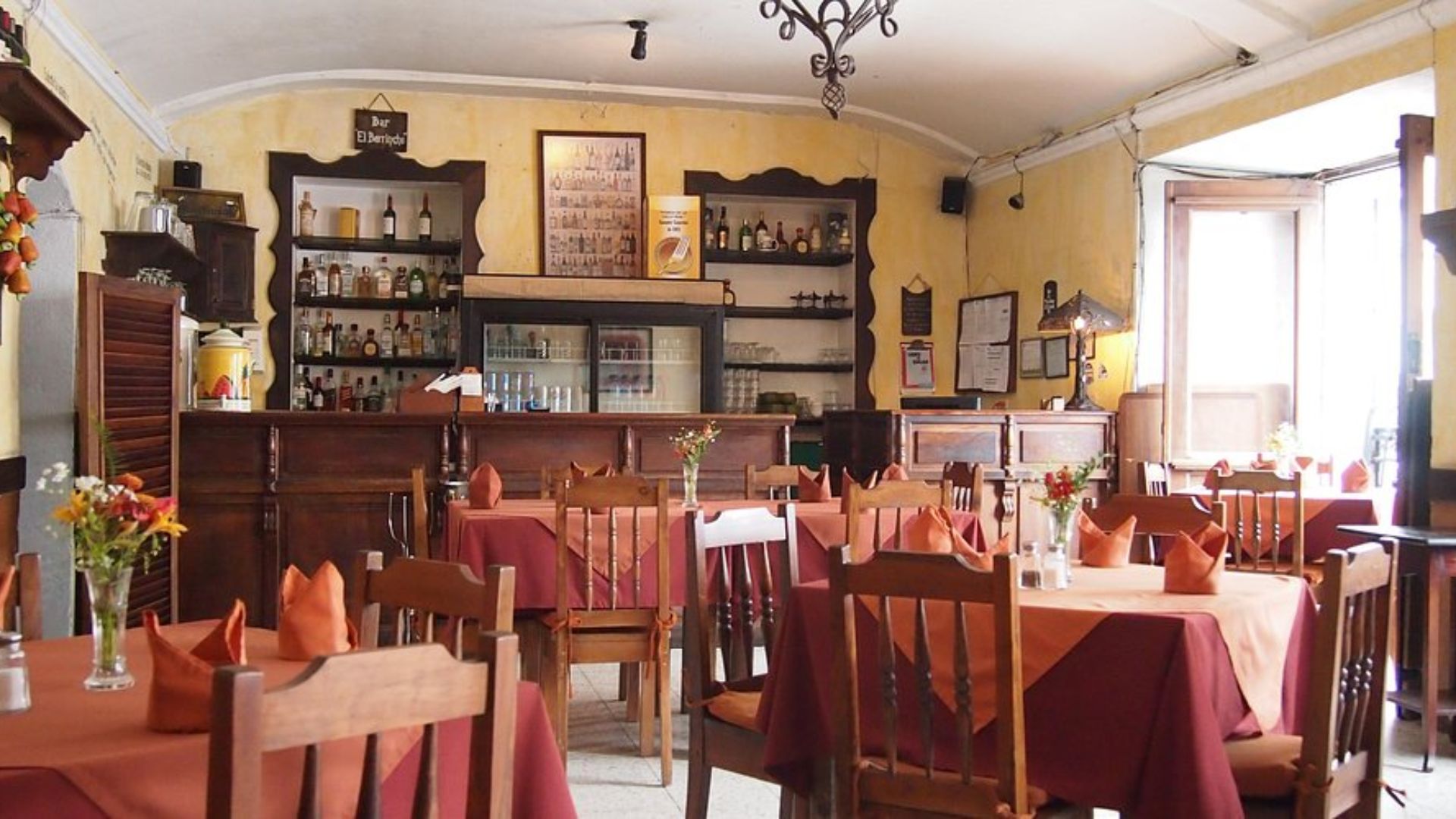 Cozy restaurant with warm lighting, wooden chairs, and tables draped in red tablecloths. A wooden bar in the background is stocked with bottles.
