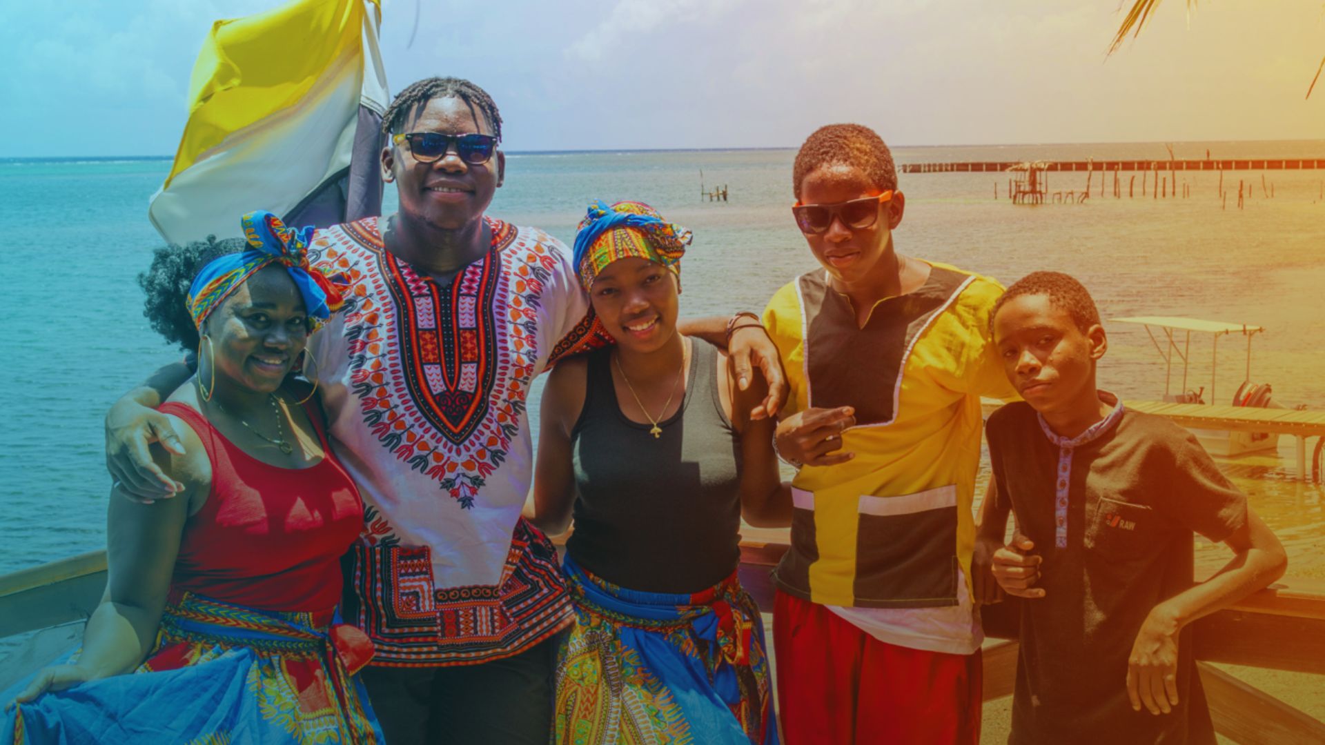 A group of five people smiling by the ocean. They are wearing colorful, traditional outfits. The setting is bright and sunny, conveying joy and warmth.