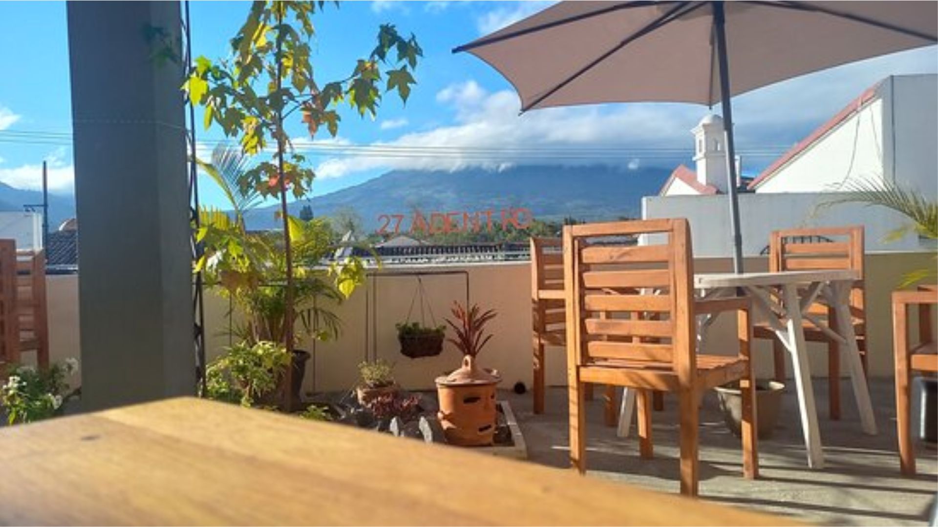 Outdoor patio with wooden tables and chairs under a large umbrella. A mountain range is visible in the background, framed by a clear blue sky and a few clouds, creating a serene atmosphere. Lush greenery and potted plants add a touch of nature.