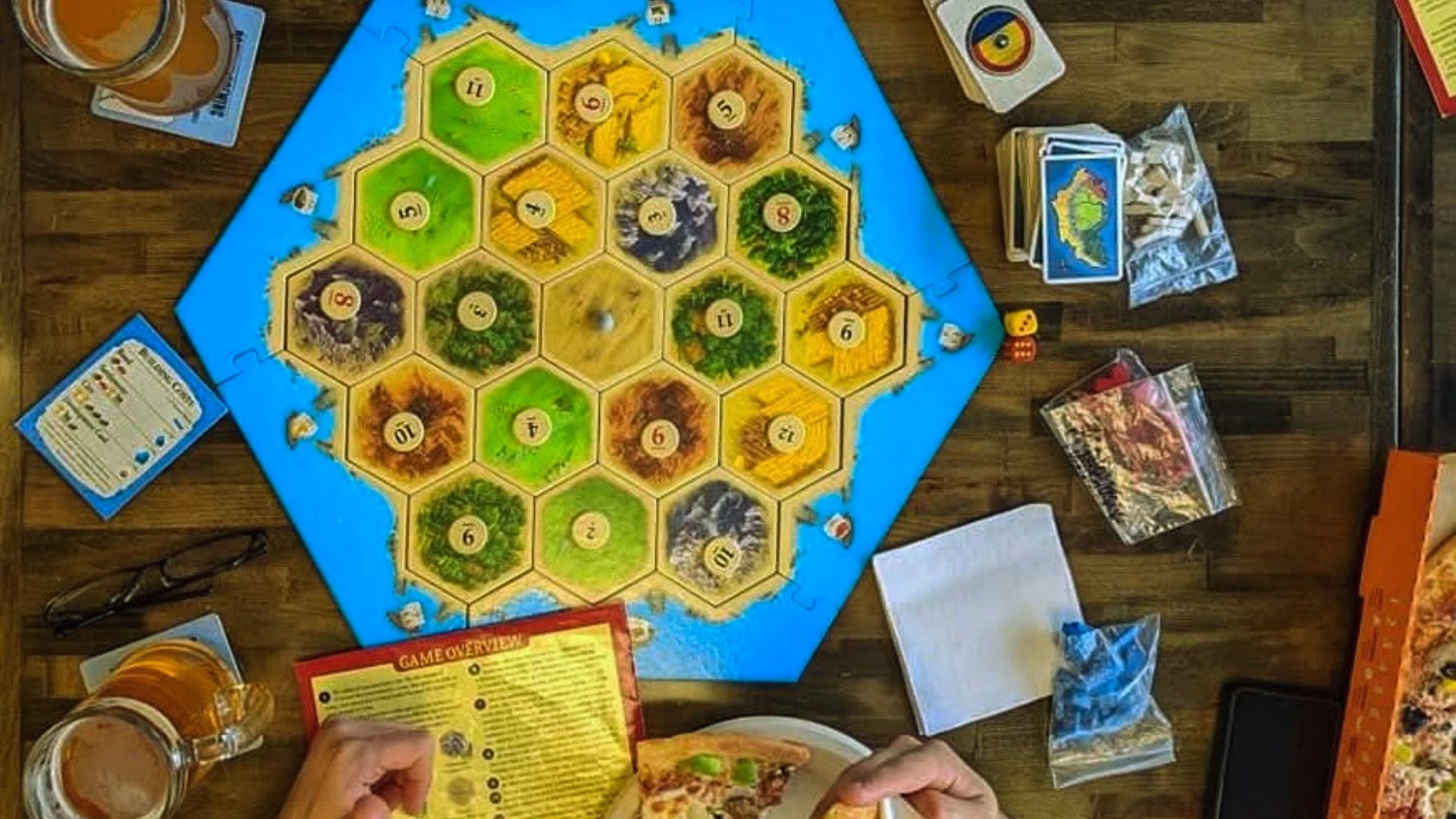 A Catan board game setup on a wooden table, surrounded by cards, dice, drinks, and pizza. Hands are seen holding a pizza slice, indicating a casual, fun atmosphere.
