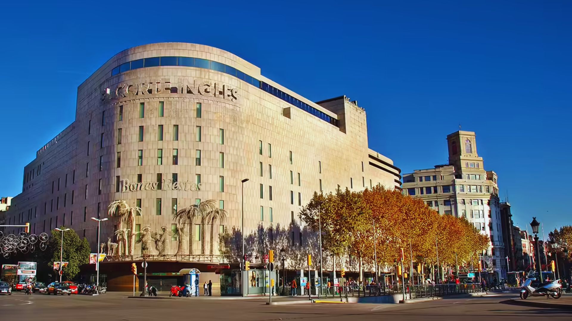 Large, modern building with "El Corte Inglés" signage, surrounded by autumn trees and a busy street under a clear blue sky, conveying a lively urban atmosphere.