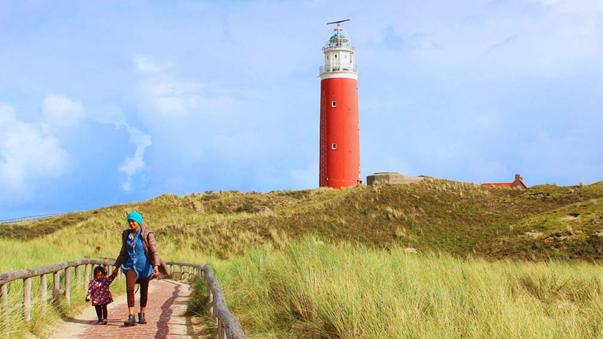 A woman and child walk on a path through grassy dunes towards a tall red lighthouse under a clear blue sky, evoking a sense of adventure and tranquility.