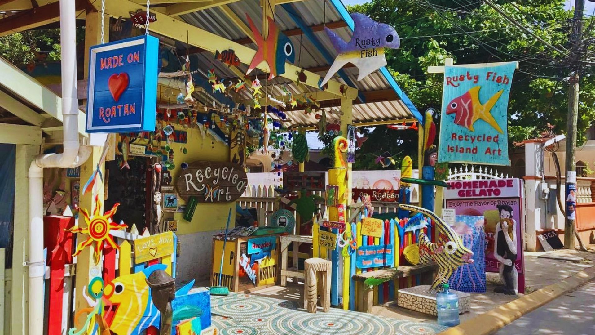A vibrant outdoor market stall displays colorful recycled art, including fish and sun motifs. Signs read "Made on Roatan" and "Recycled Island Art," creating a lively, eco-friendly atmosphere.