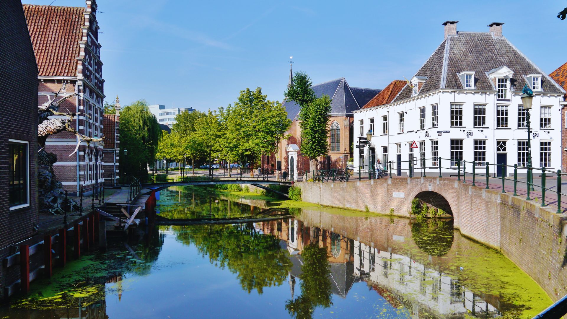 Quaint canal scene with historic brick buildings, arched bridge, and lush green trees reflected in the calm water, creating a serene atmosphere.
