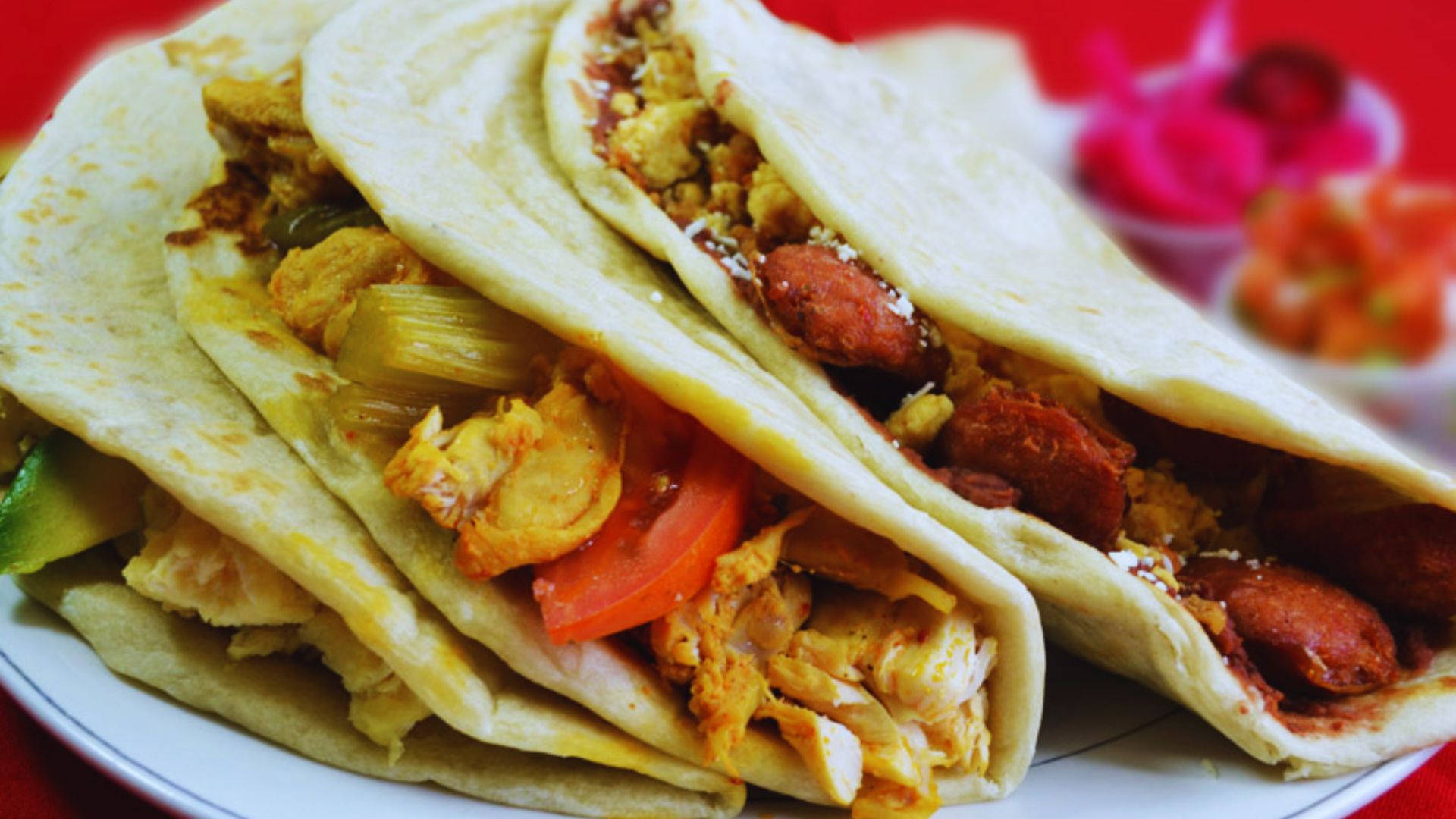 Three folded tacos filled with chicken, chorizo, and vegetables sit on a white plate. The vibrant colors and fresh ingredients convey a delicious, appetizing feel.