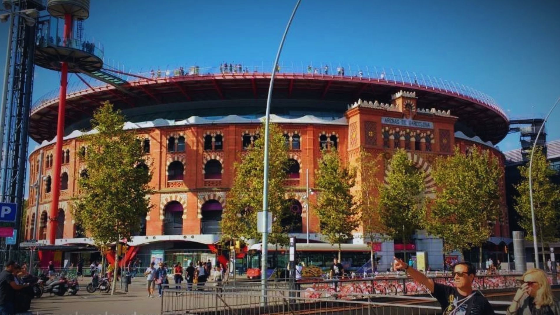 A large circular red-brick arena, adorned with arches and decorative details, is encircled by lush trees. People and vehicles bustle in the busy foreground.