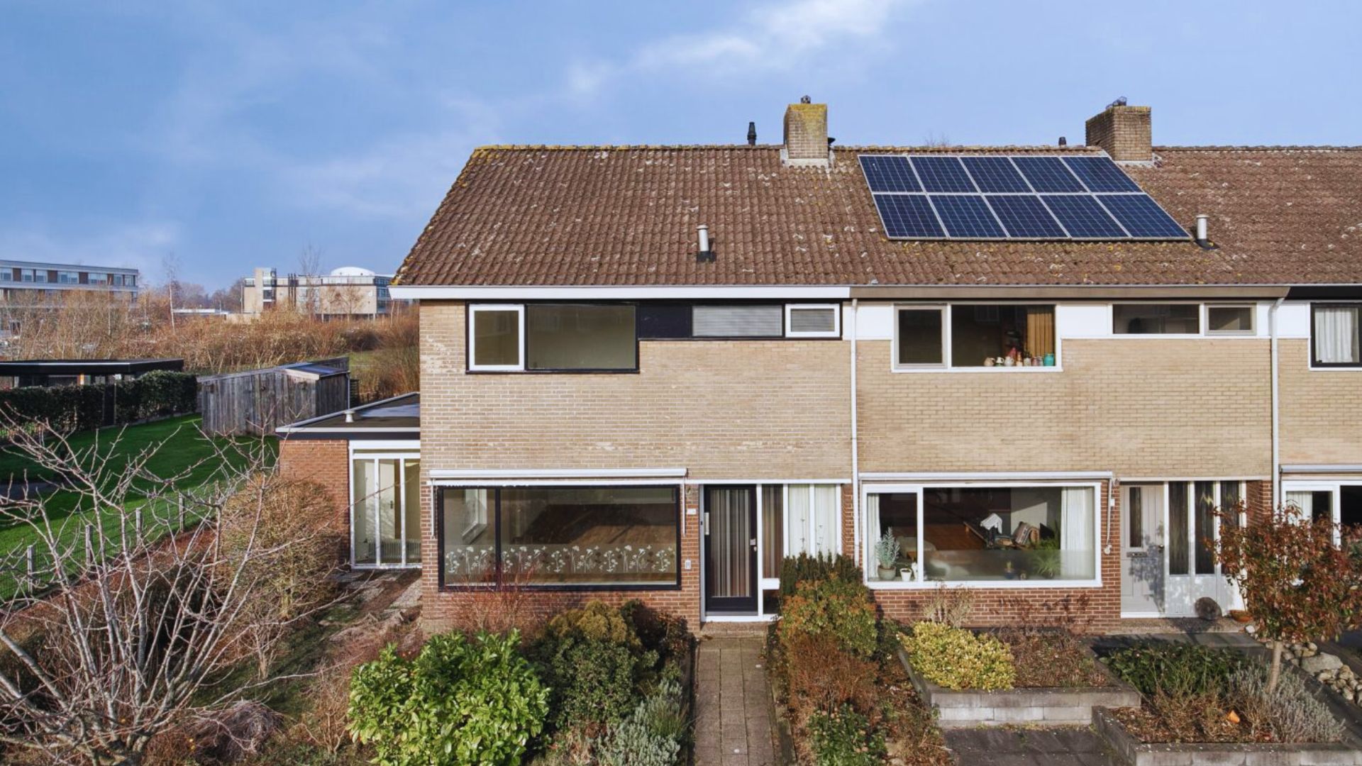 A two-story brick house with a sloped roof featuring solar panels. It has large front windows, a tidy garden, and a clear, sunny sky in the background.