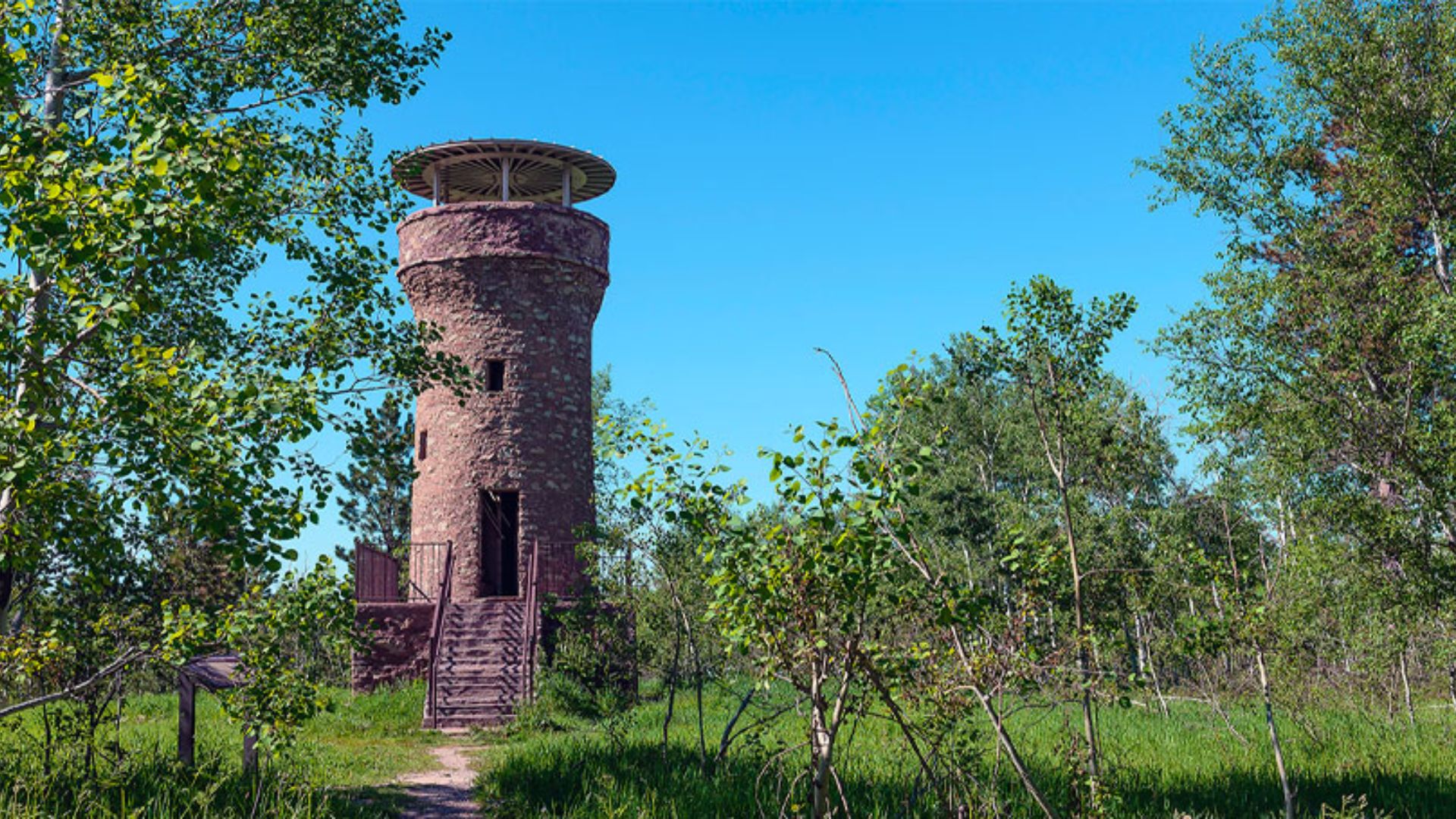 A rustic stone tower stands in a lush green forest under a bright blue sky. A narrow path leads to its entrance, evoking a sense of exploration and tranquility.