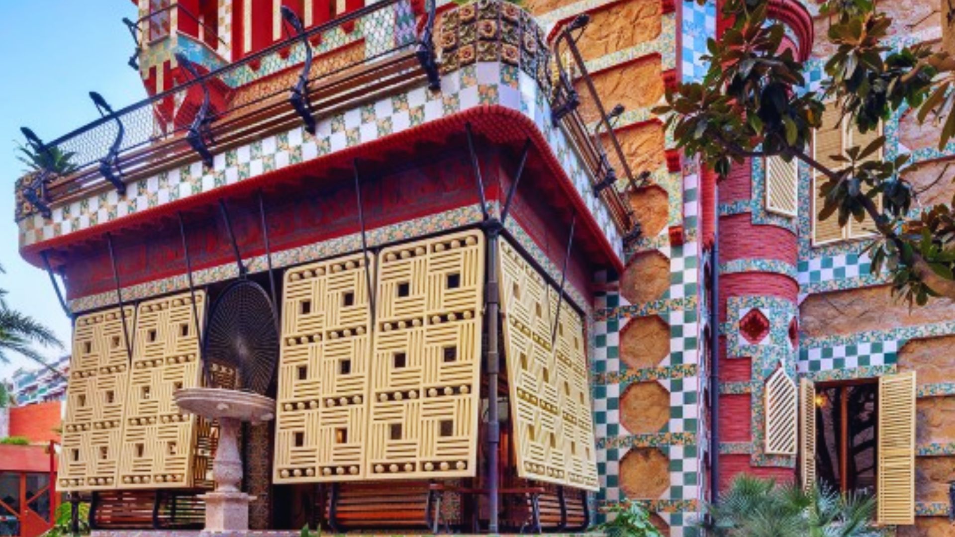 Colorful building facade with intricate geometric patterns, featuring a blend of red, yellow, and green tiles. Decorative balcony and lush greenery.