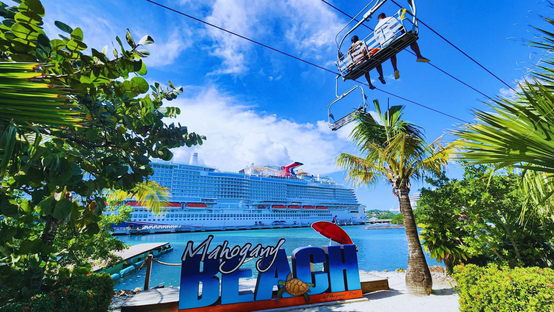 A cruise ship docked at Mahogany Beach on a sunny day. Lush green palm trees and a vibrant "Mahogany Beach" sign are in the foreground. Aerial chairlift with people overhead.